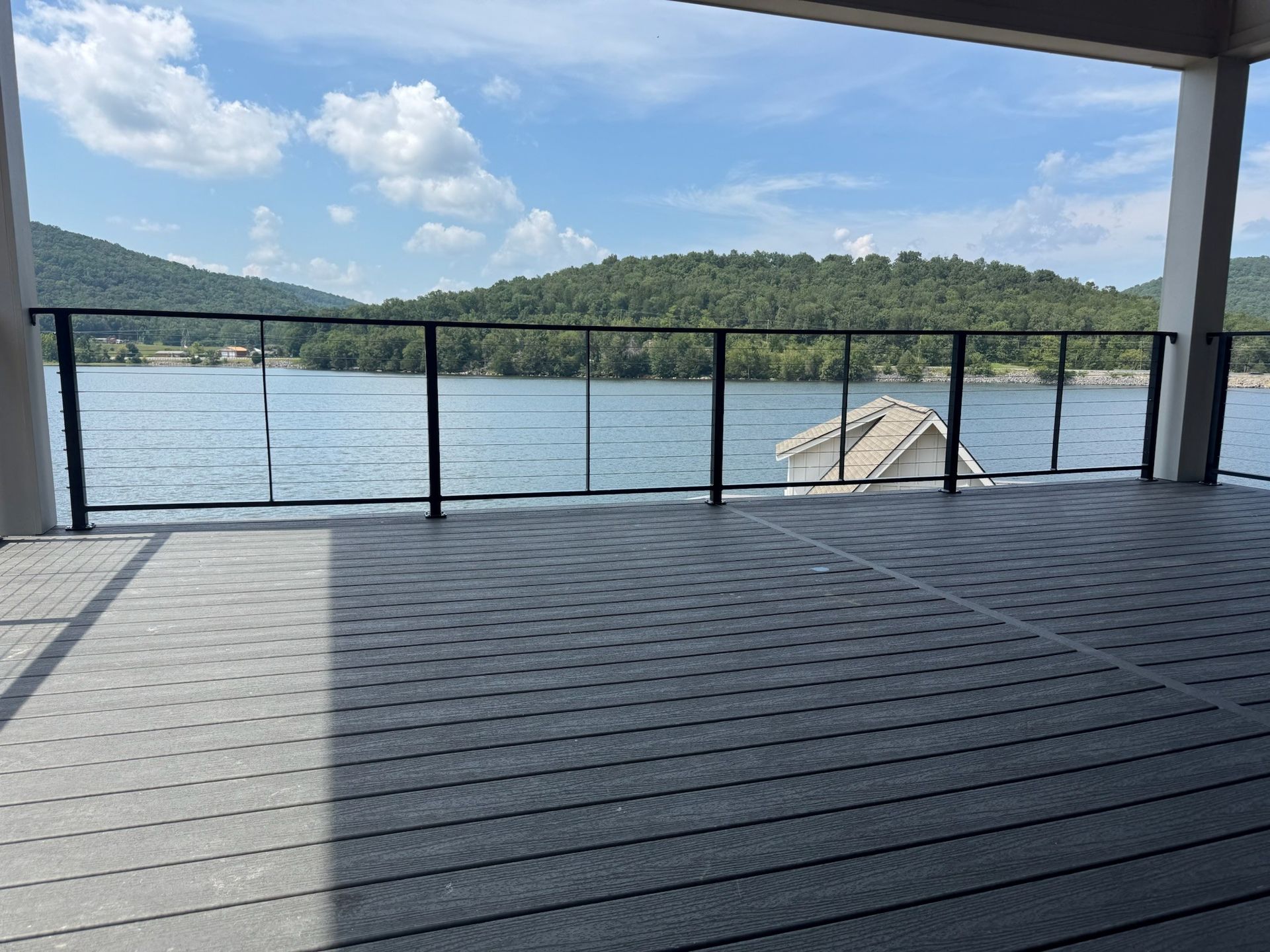 Covered lakeside deck with black railing, calm water, and green hills under a blue sky