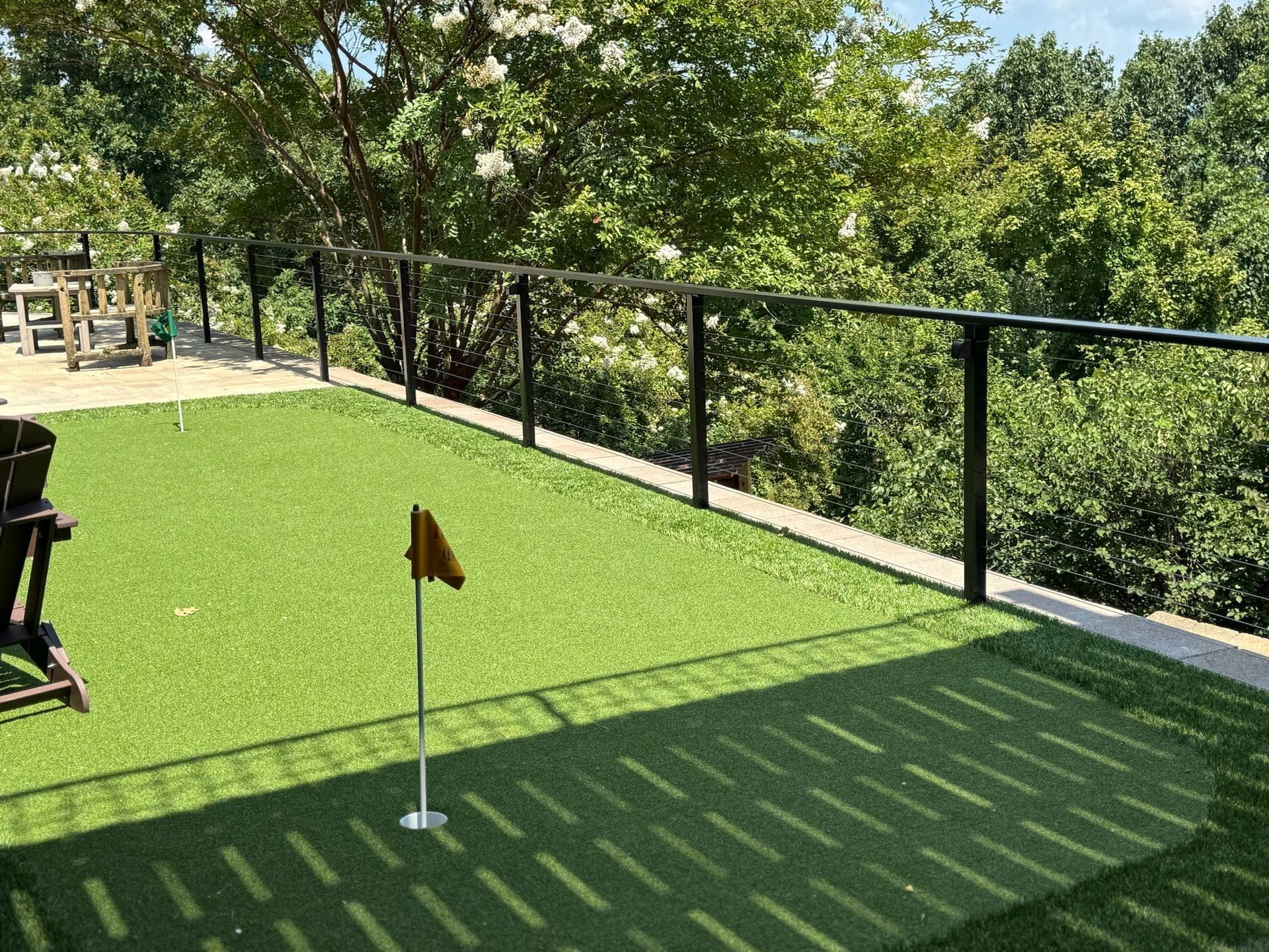 Mini-golf green with a hole flag, black fence, and trees in the background.