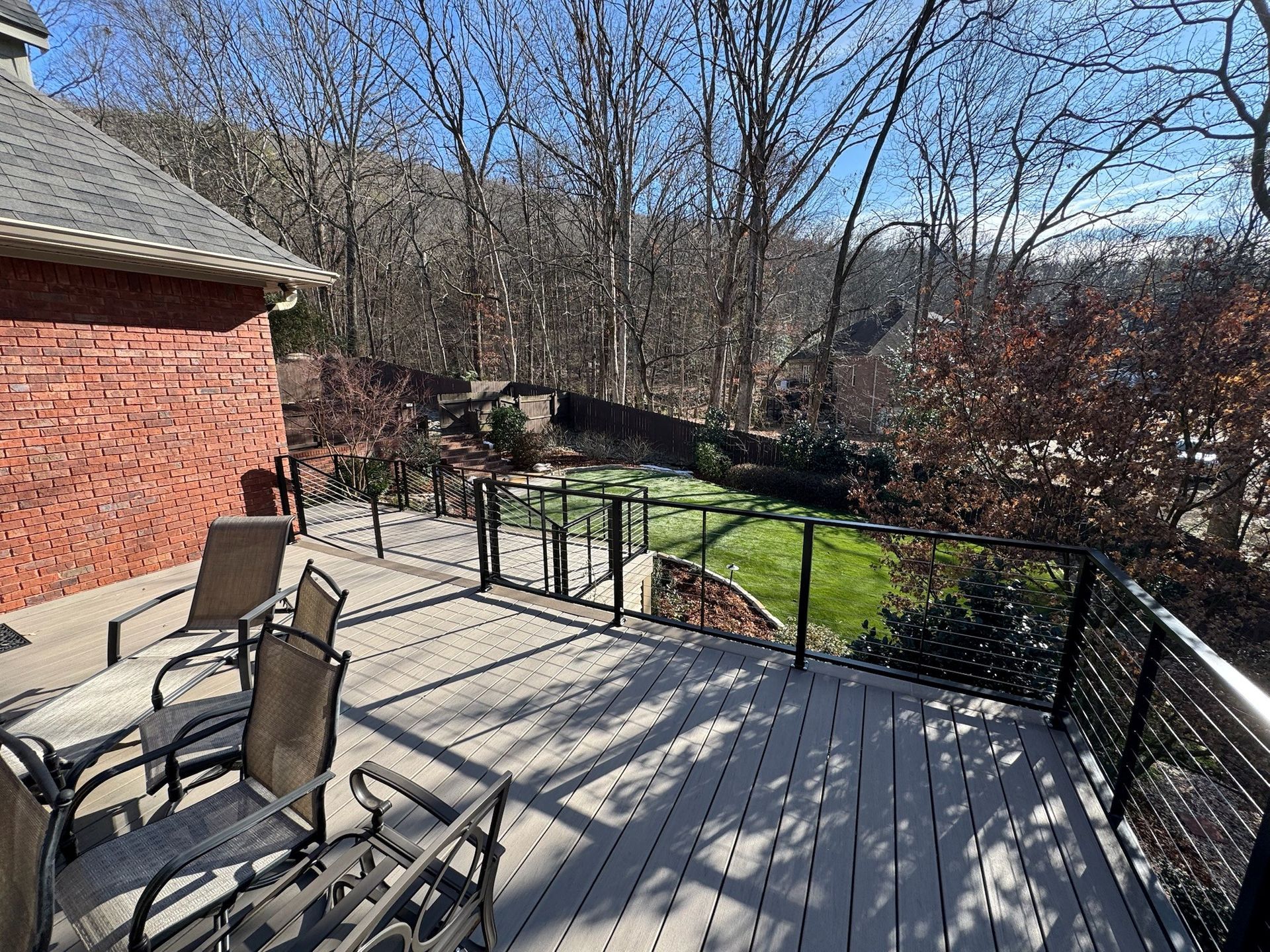 Backyard deck with patio chairs, black railing, brick wall, and leafless trees in sunlight