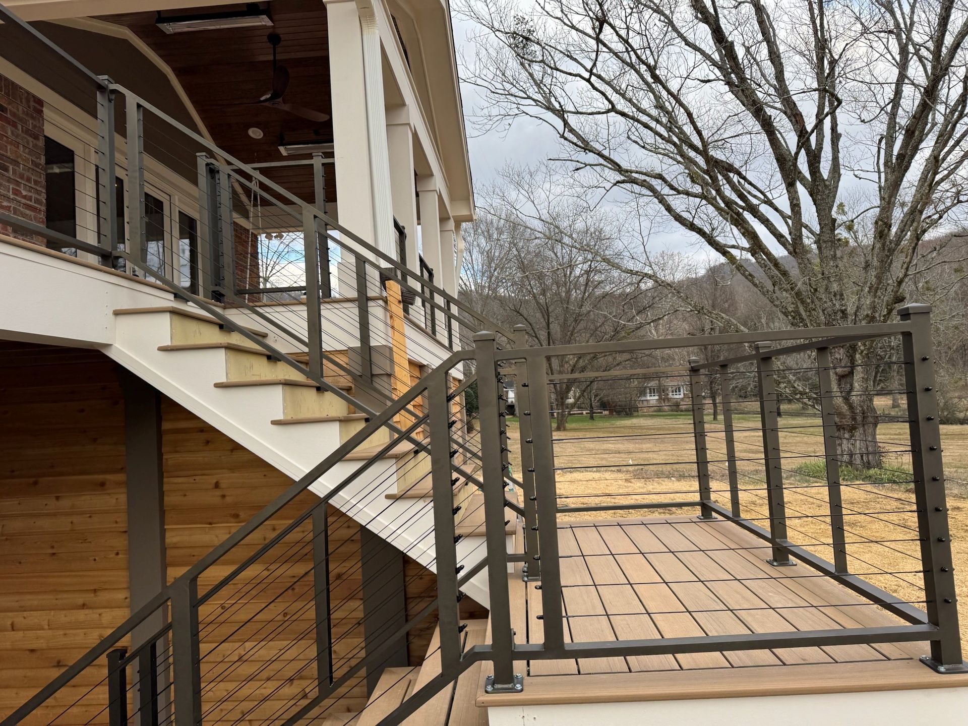 Wooden house porch with stairs and a ramp, bordered by black railings in a yard with leafless trees.