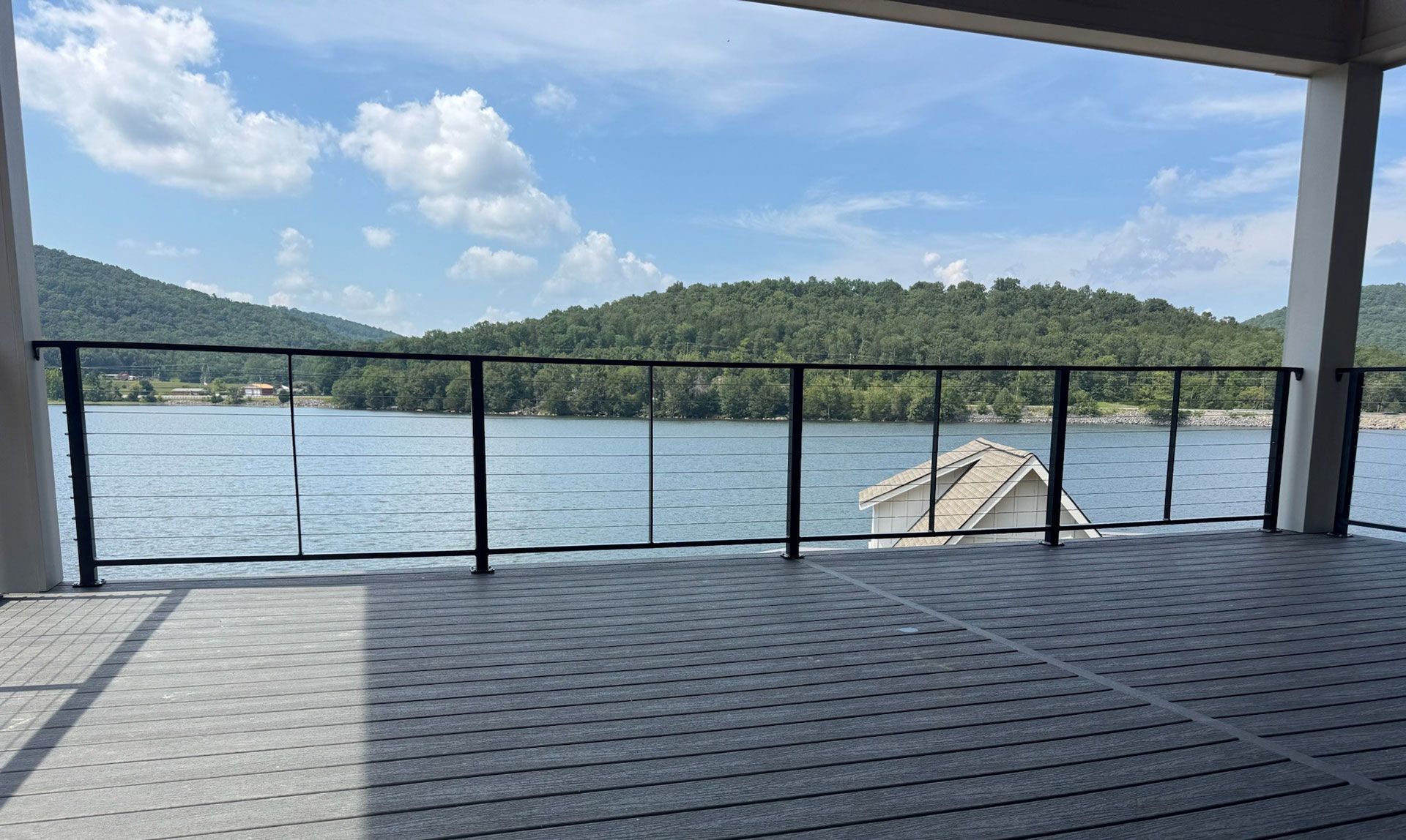 Covered lakeside deck with black railing, calm water, green hills, and a small dock under a blue sky