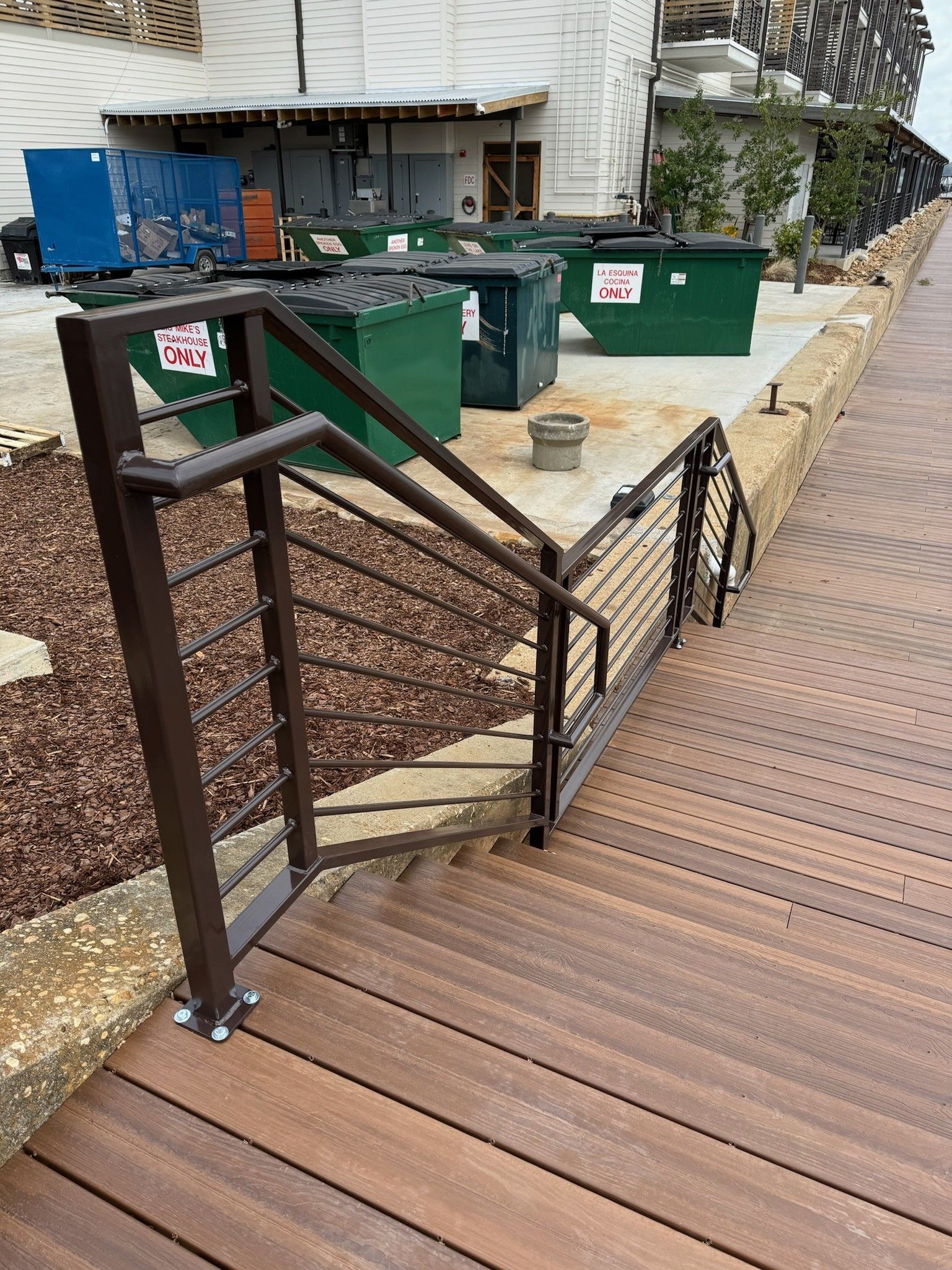 Wooden boardwalk ramp with black handrails beside green trash bins and a building wall