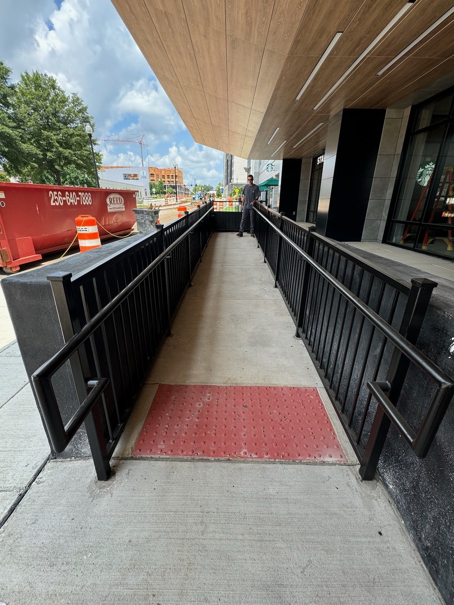 Accessible ramp with black railings, red tactile mat, and overhead awning beside a building.