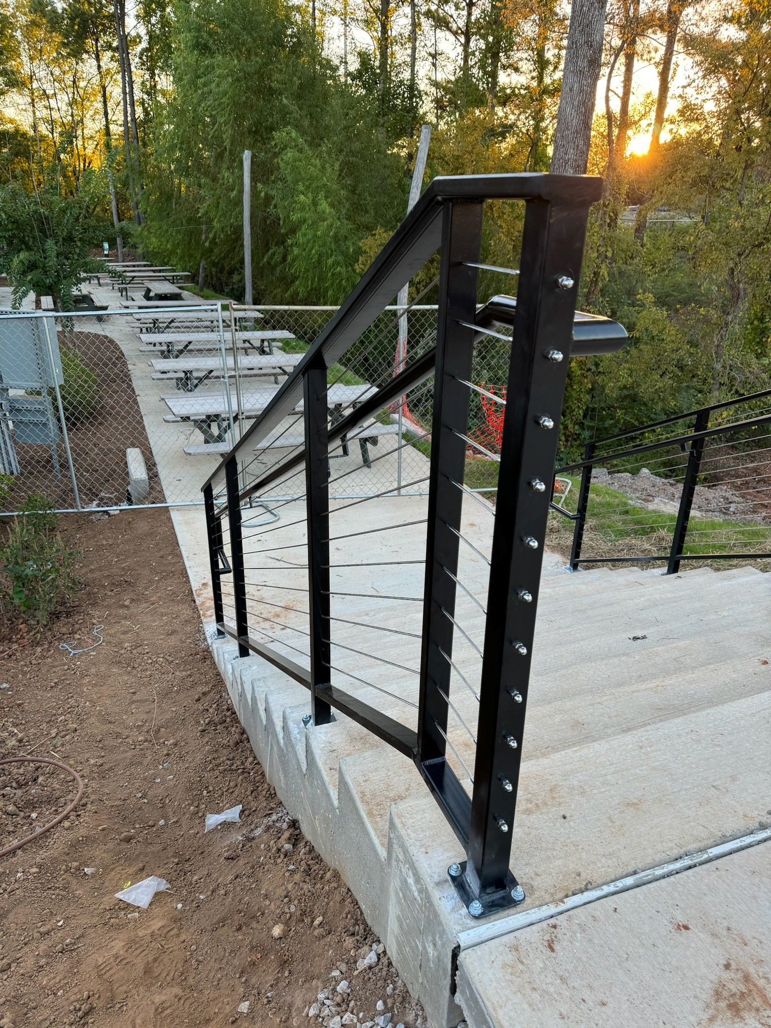 Black metal railing along curved concrete steps outdoors at sunset with trees and gravel around it