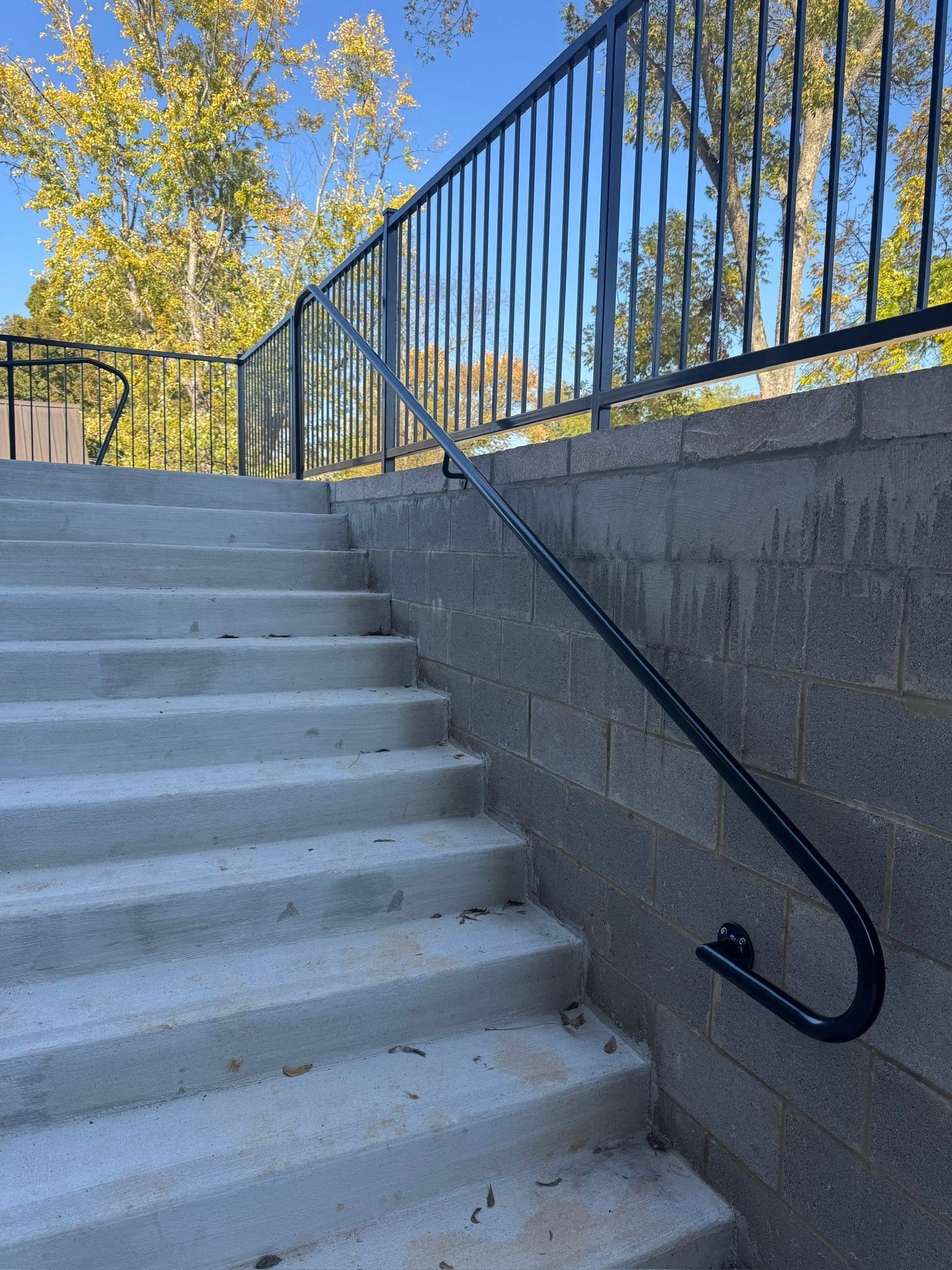 Concrete outdoor stairway with black metal handrails beside a stone wall under blue sky.