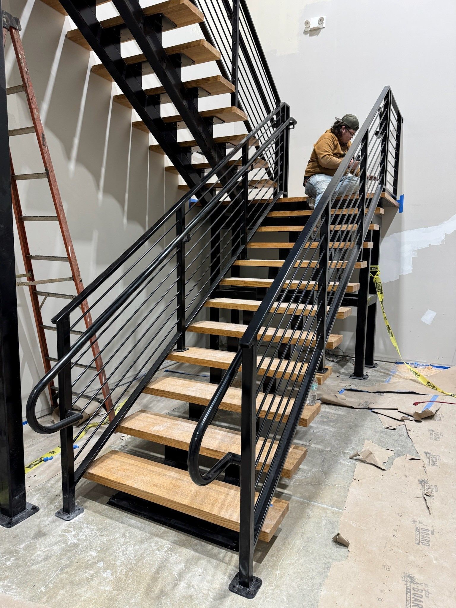 Black metal staircase under construction, with a worker near the top and tools on the floor.