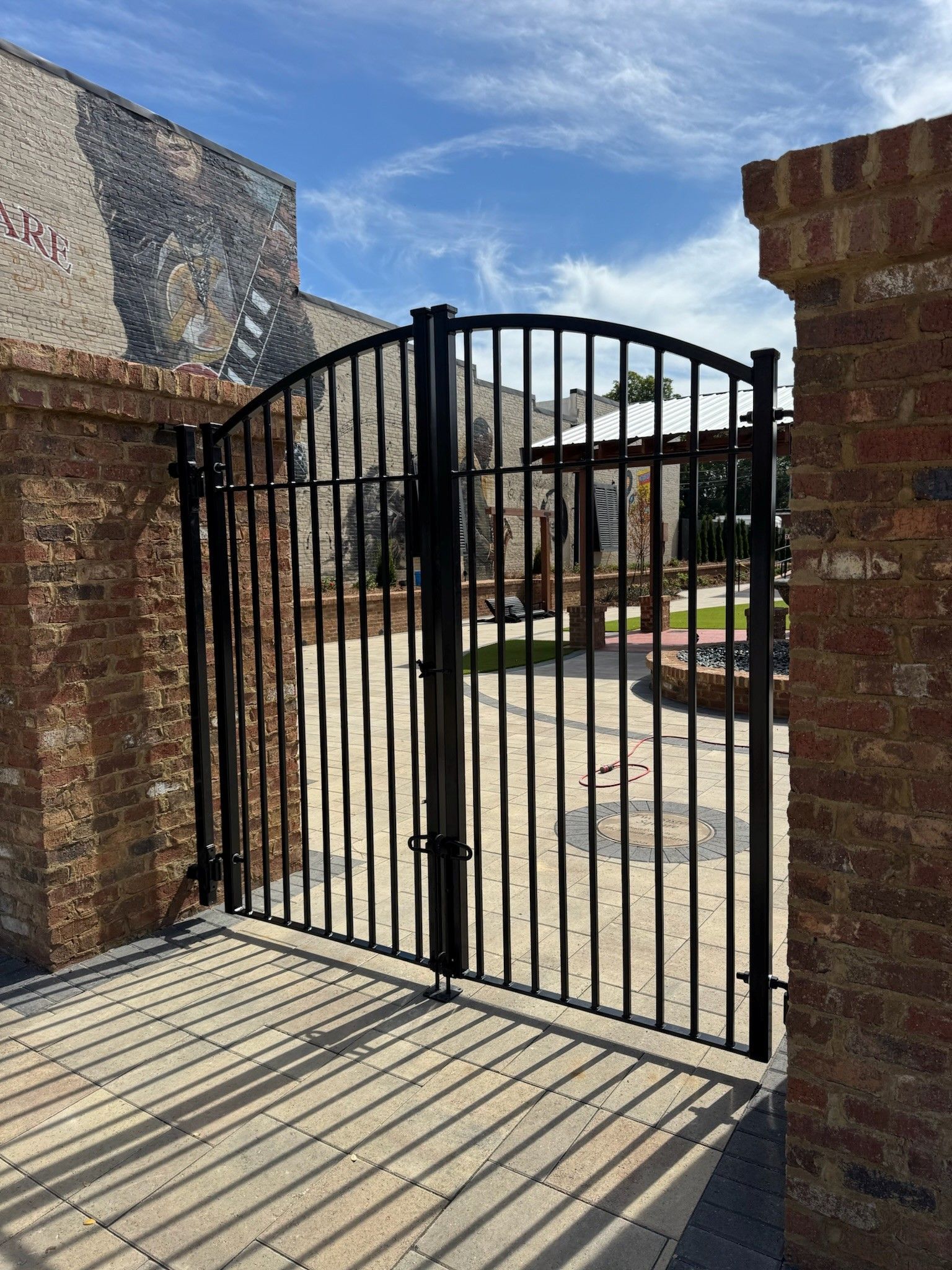 Closed black iron gate between brick pillars, opening to a sunny courtyard under a blue sky