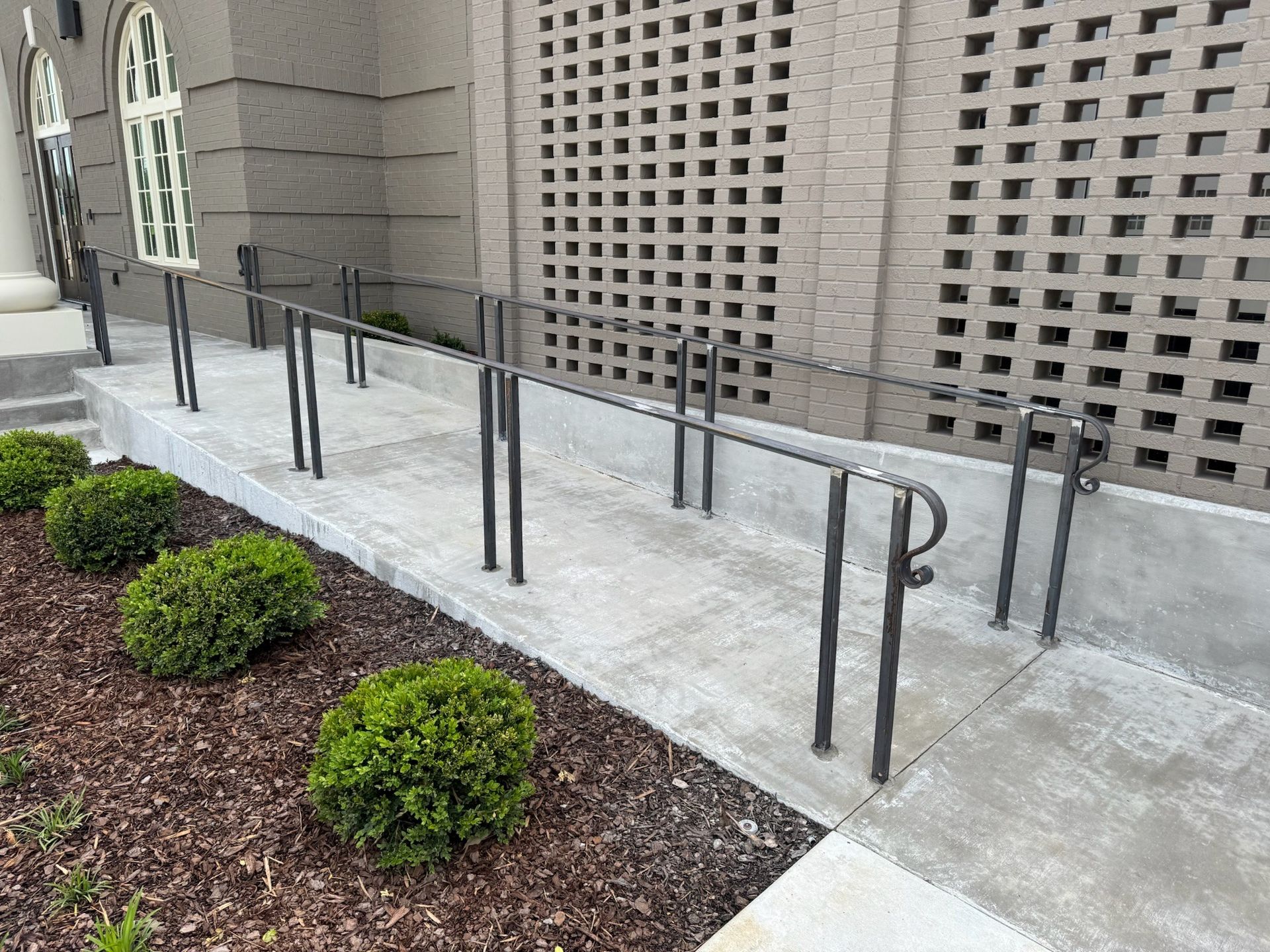 Outdoor concrete ramp with black metal handrails beside a beige brick building and small shrubs.