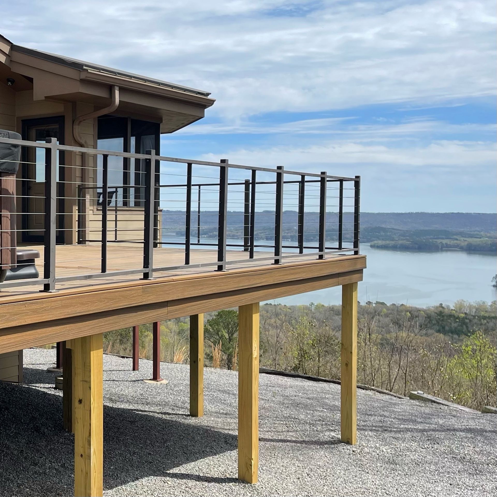 Wooden deck with black railings beside a house, overlooking a lake and distant hills under a cloudy sky