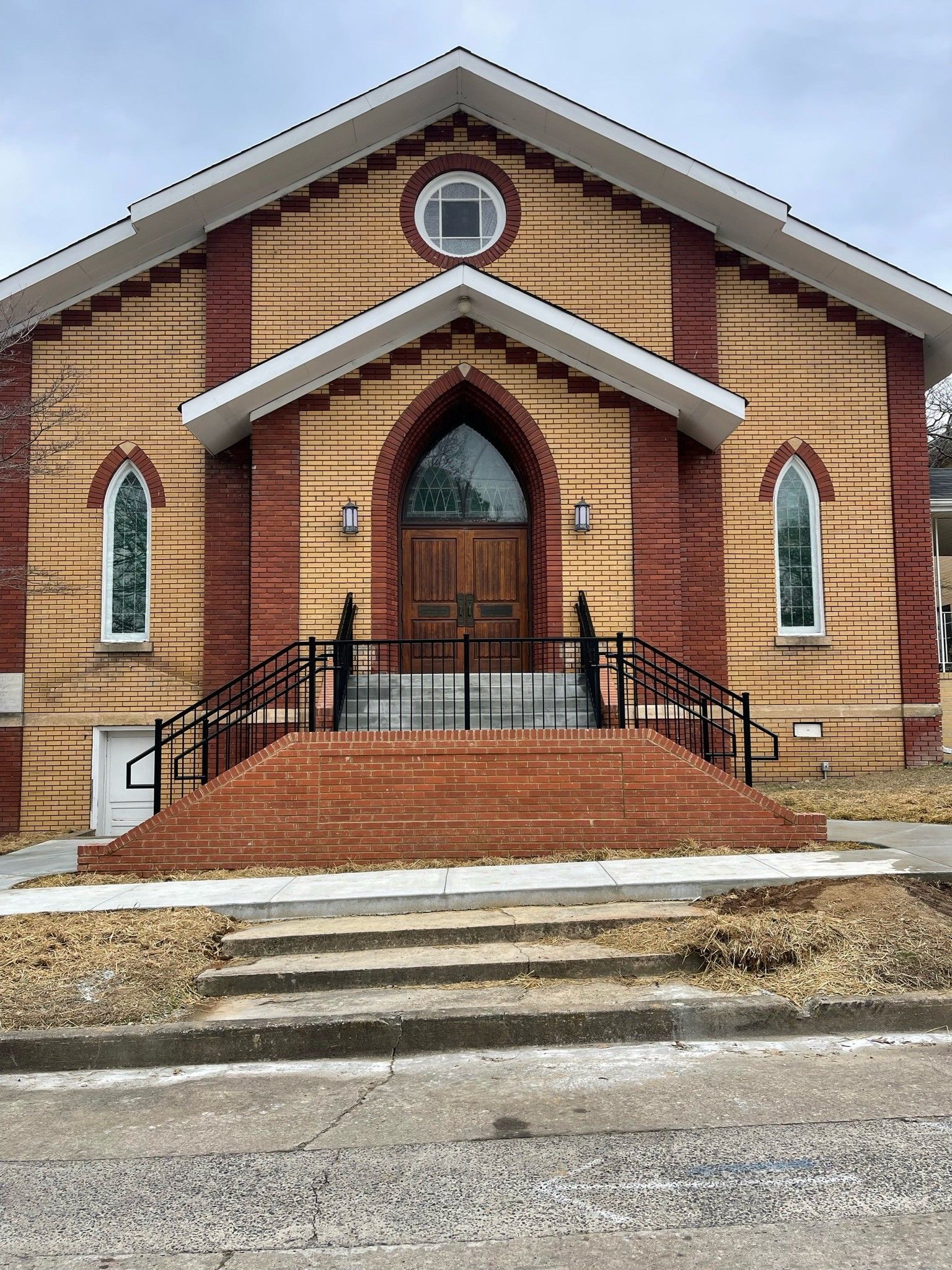 Front view of a small brick church with arched windows, a central doorway, and steps leading up from the street.