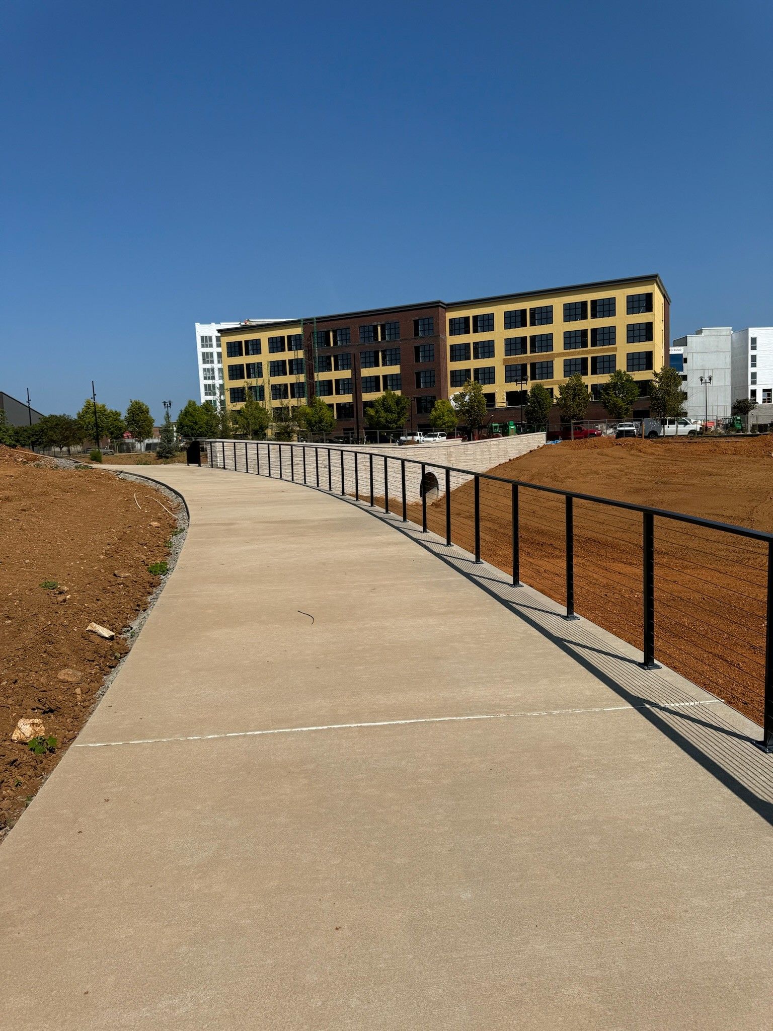 Curved concrete path beside a fenced dirt lot, leading toward a yellow apartment building under a blue sky.
