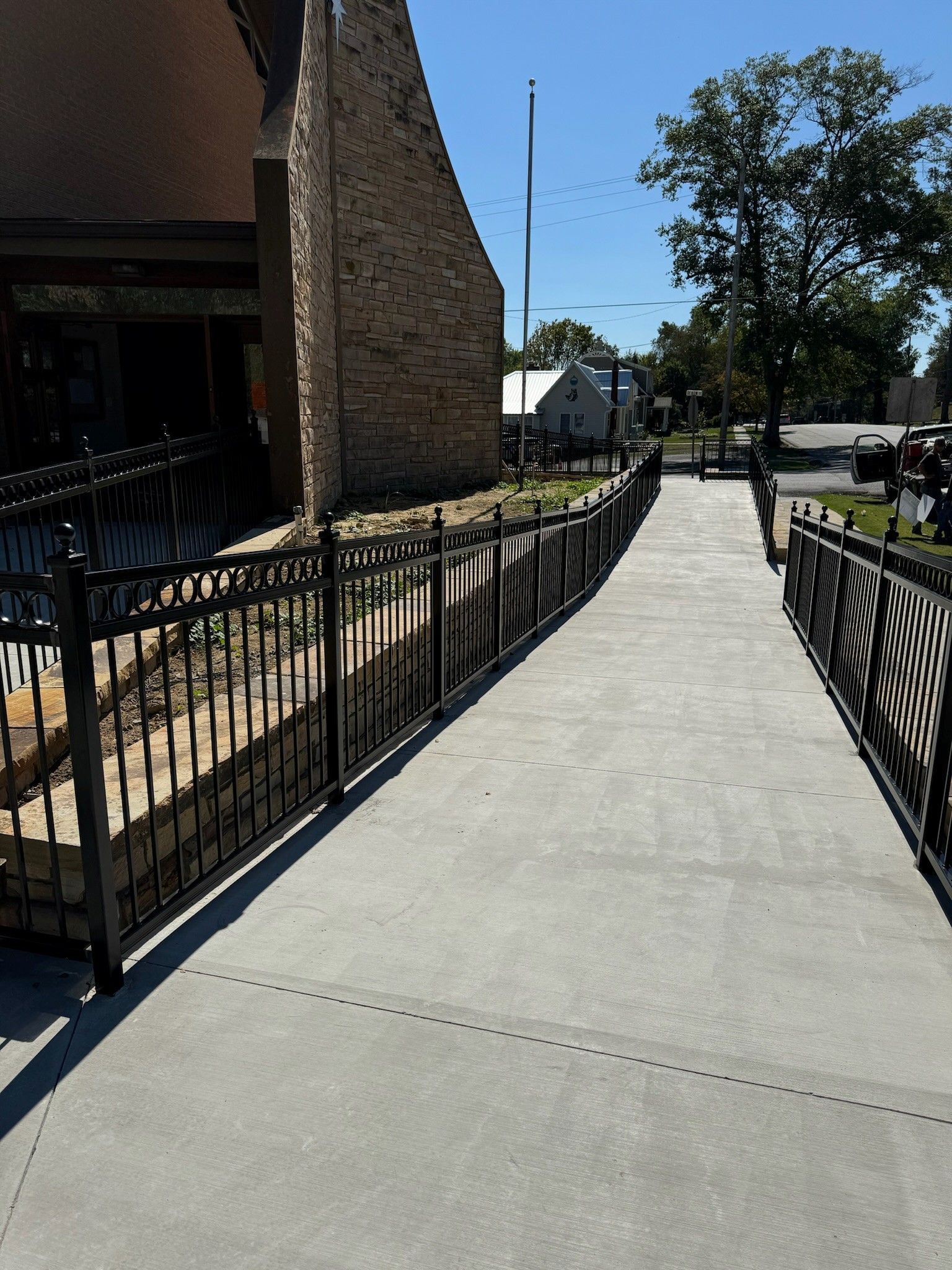 Sunlit concrete ramp with black railings beside a stone building and street trees