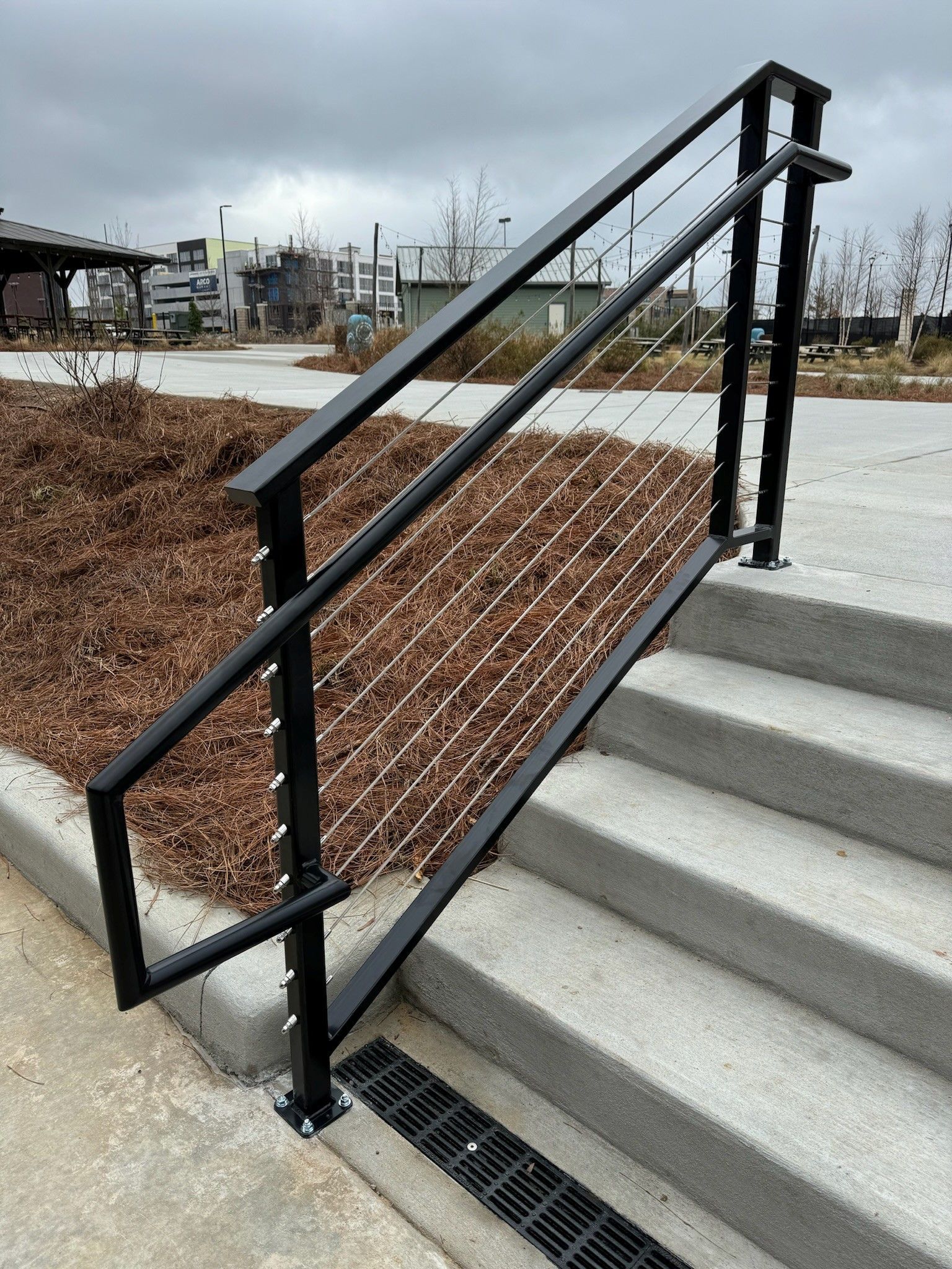 Black handrail and mesh ramp beside outdoor concrete steps, with a drain at the bottom.