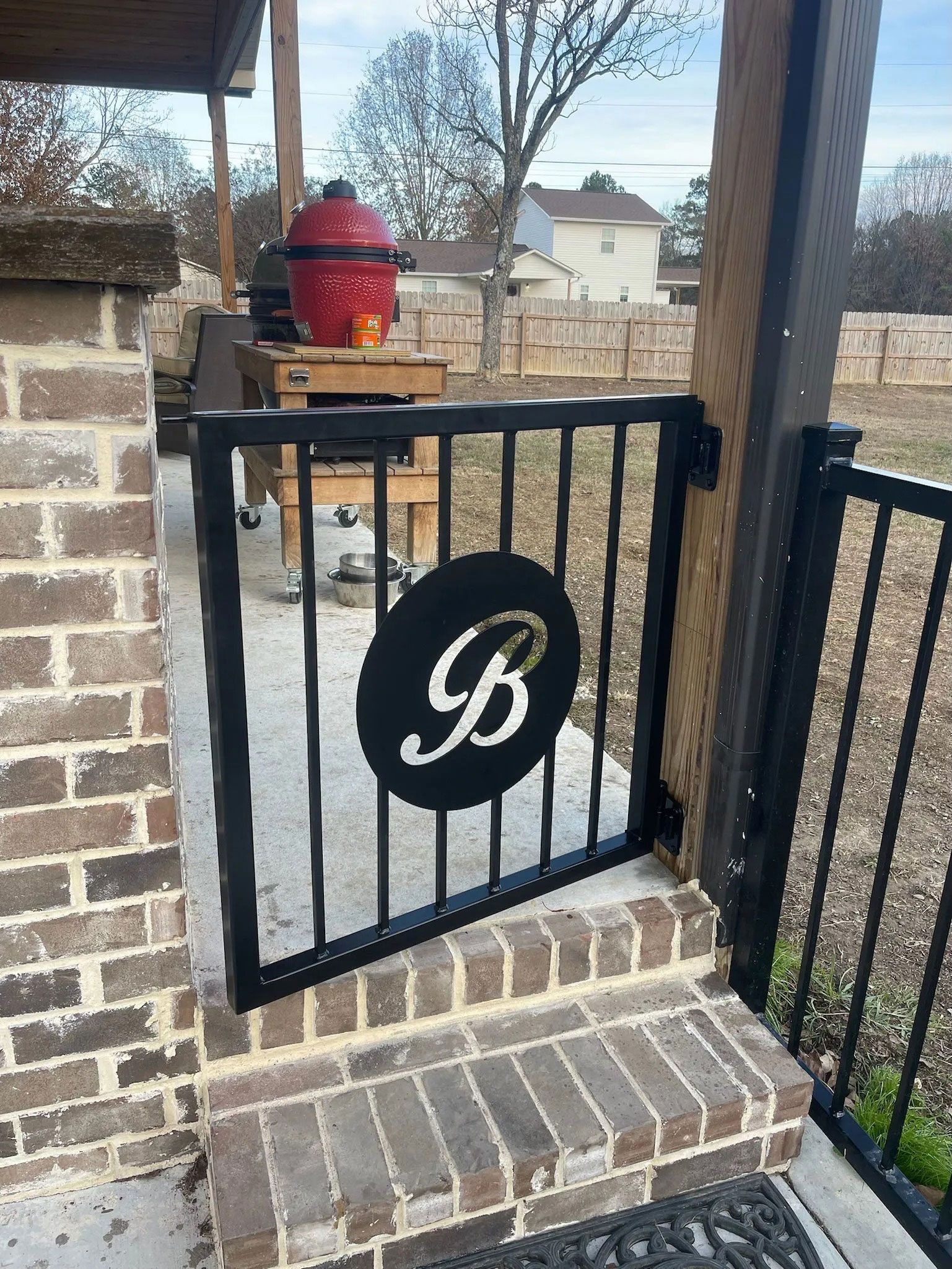 Black metal gate with a white monogram beside brick steps and a red planter on a small table outside a home