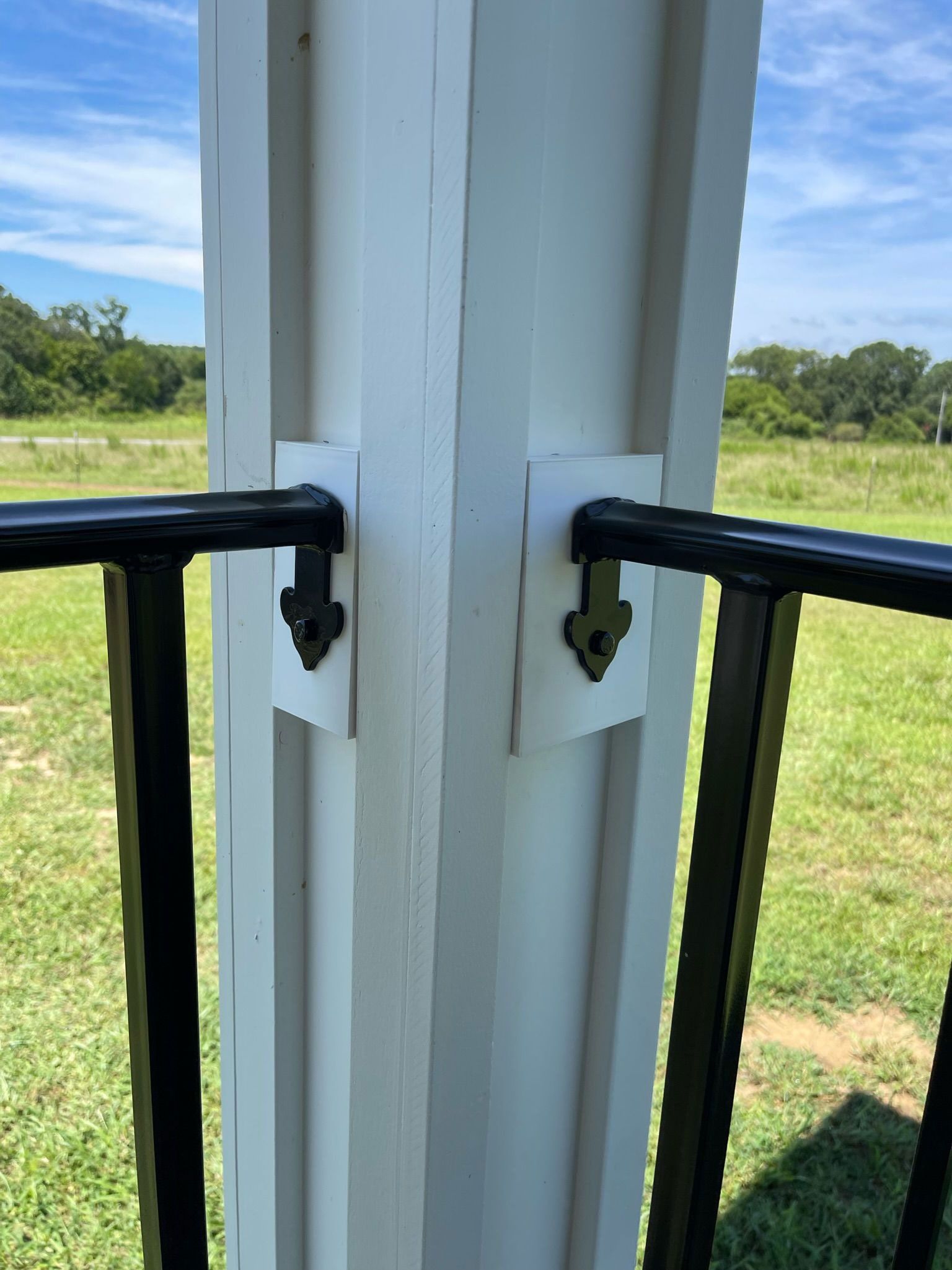 Black handrails attached to a white porch post, overlooking a green field under a blue sky.