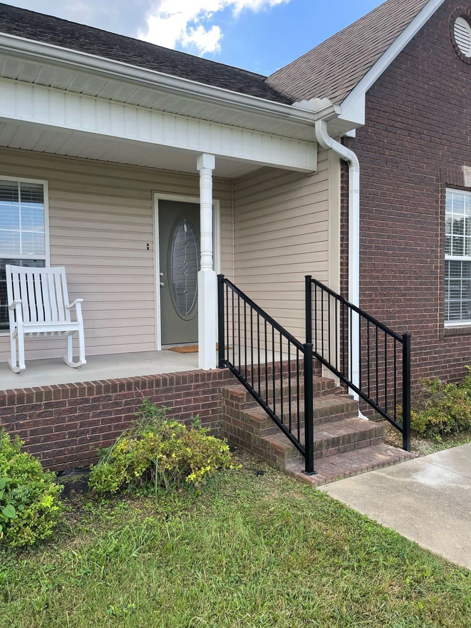 Front porch with white chair, black railing, and brick steps beside a beige house.