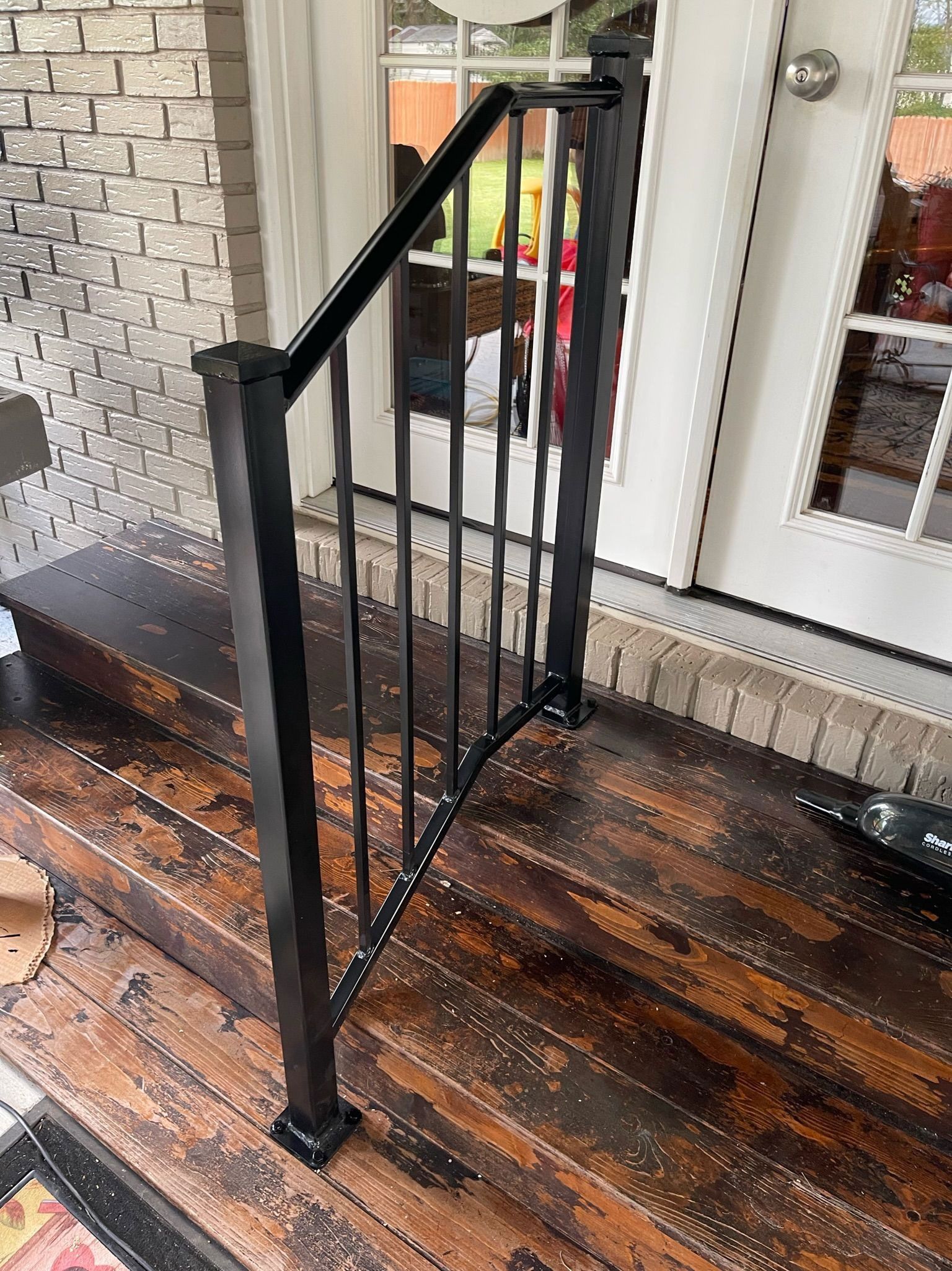 Black metal railing on a wet wooden porch beside a white door and brick wall.