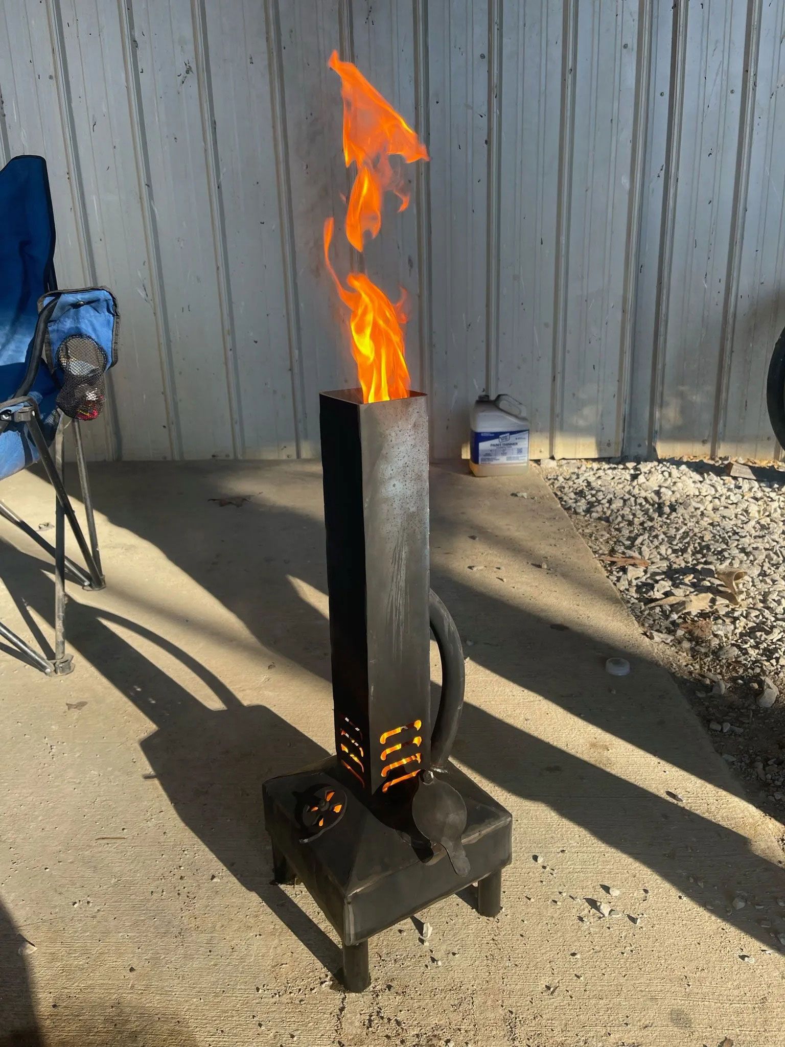 Metal fire pit with tall orange flames burning outdoors on a concrete patio.