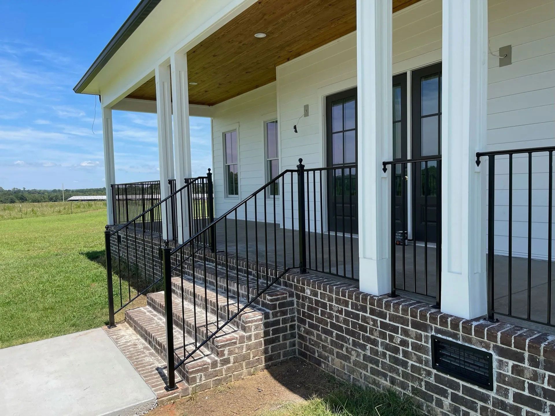 Front porch with black railings and brick steps on a white house overlooking a grassy field