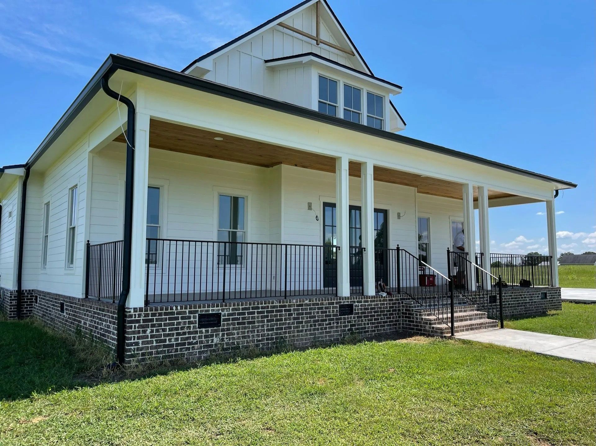 White house with a wraparound porch, black railings, and a grassy yard under a clear blue sky