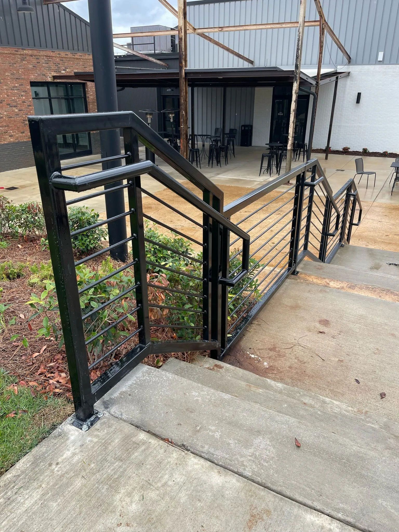 Black metal railing borders a sloped concrete walkway beside a brick patio and patio furniture