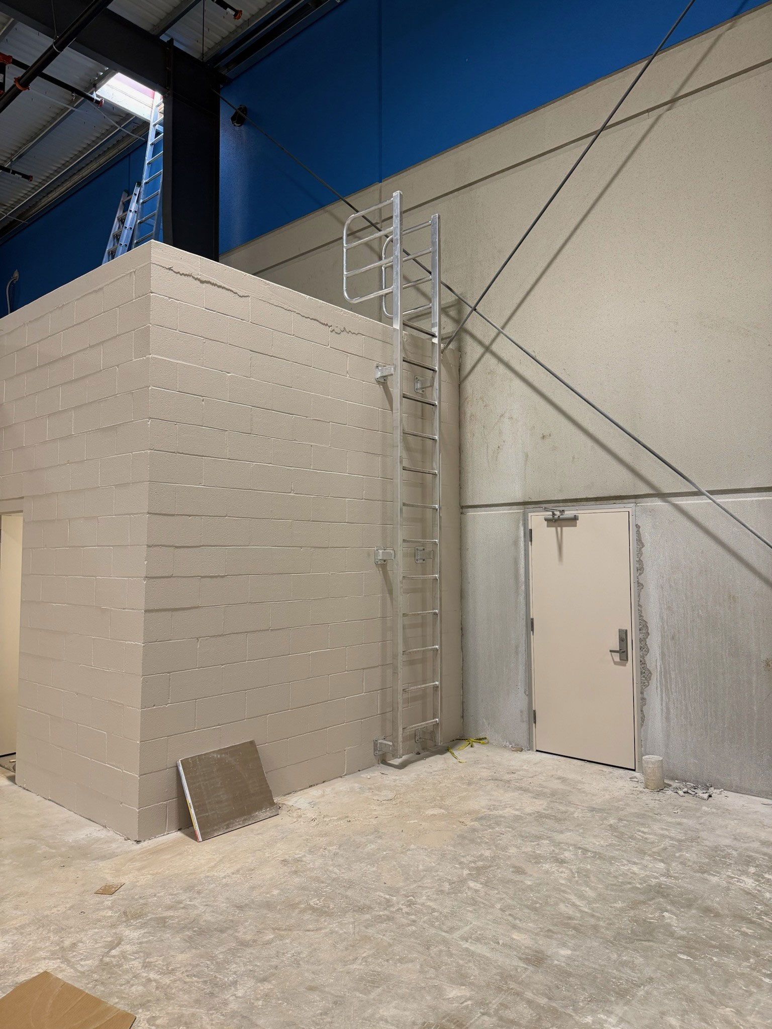 Indoor industrial space with a ladder beside a white brick wall and a gray door, blue wall above.