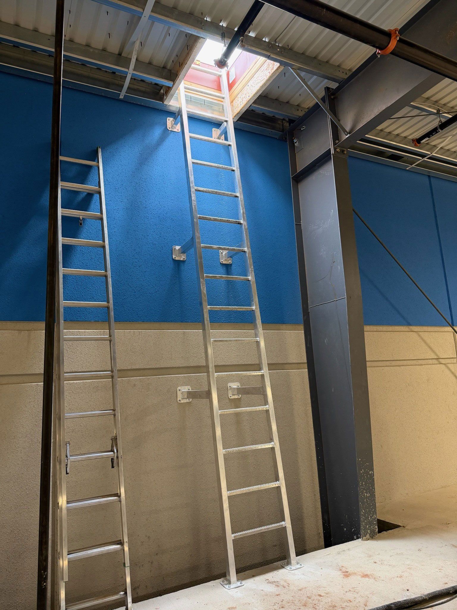 Tall ladders inside a warehouse, reaching up to a ceiling hatch above a blue wall.