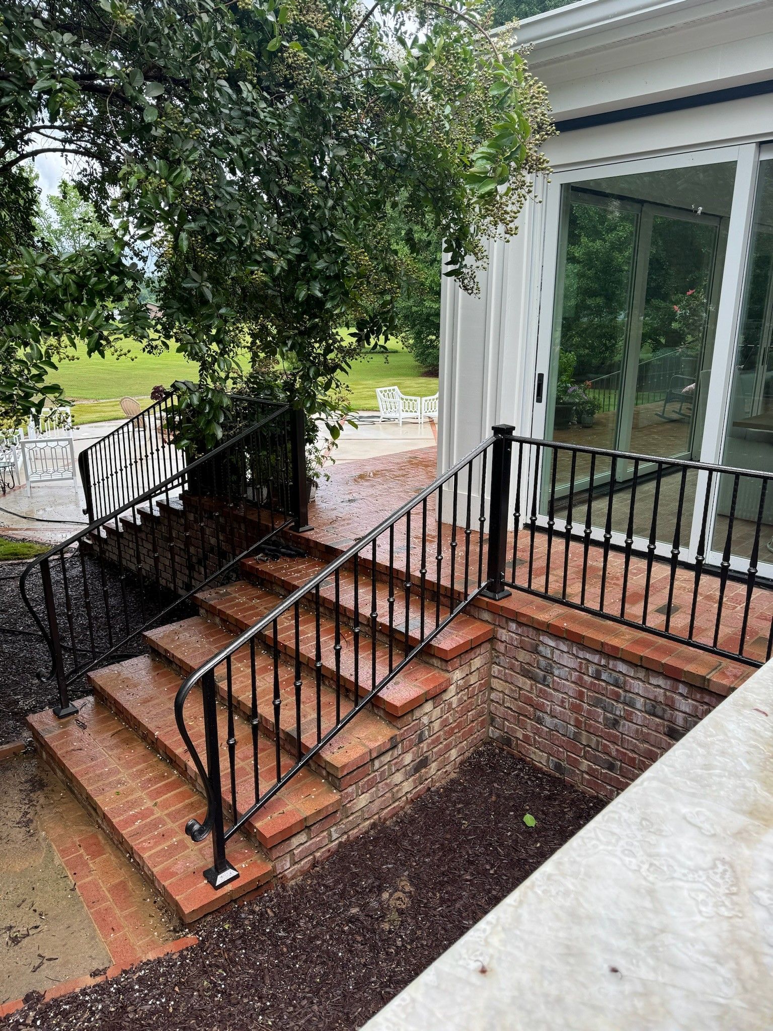 Front steps with black metal railing leading to a house entrance beside a tree and sliding glass door