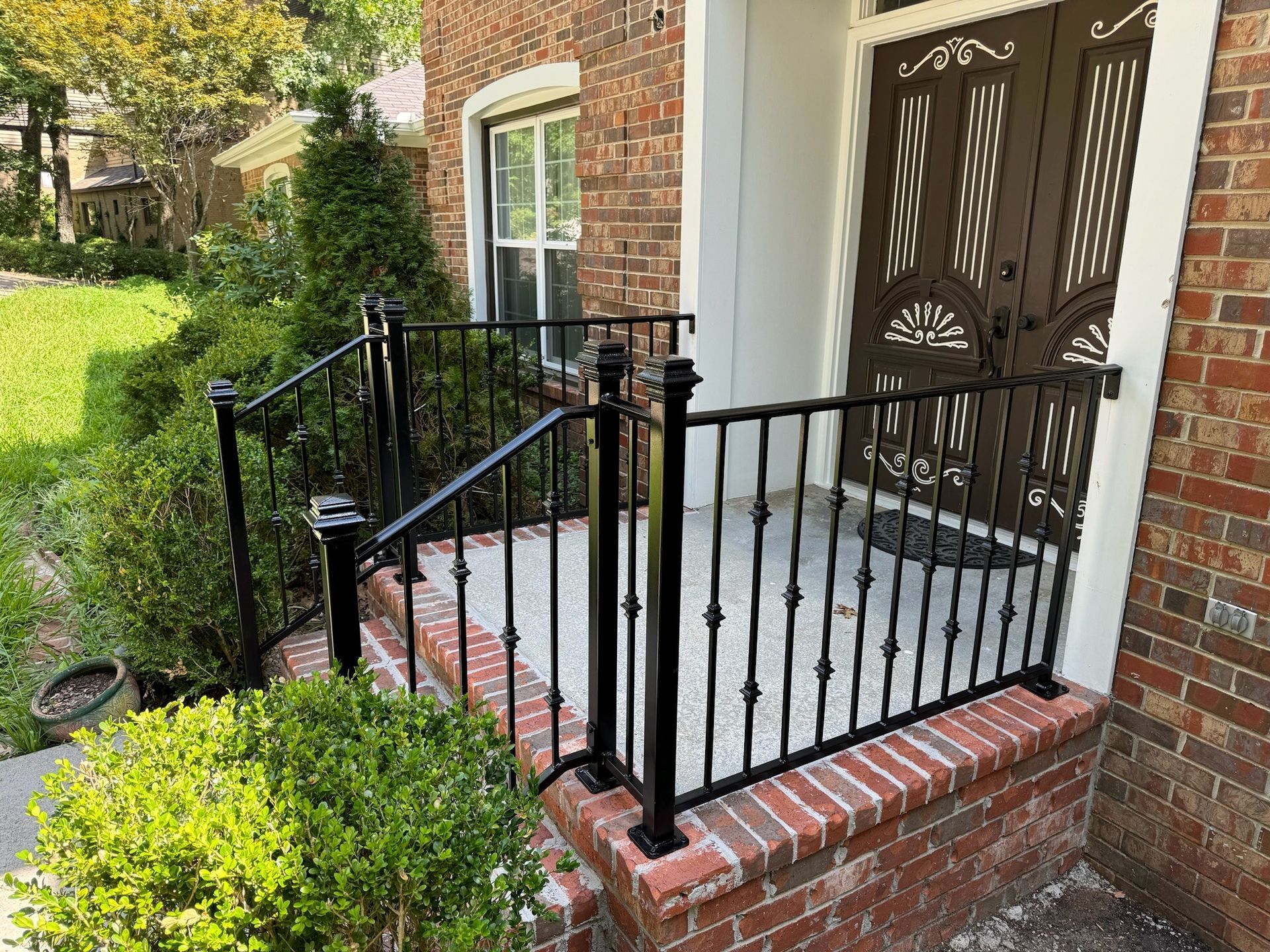 Brick front porch with black railings and steps beside a brick house and green shrubs