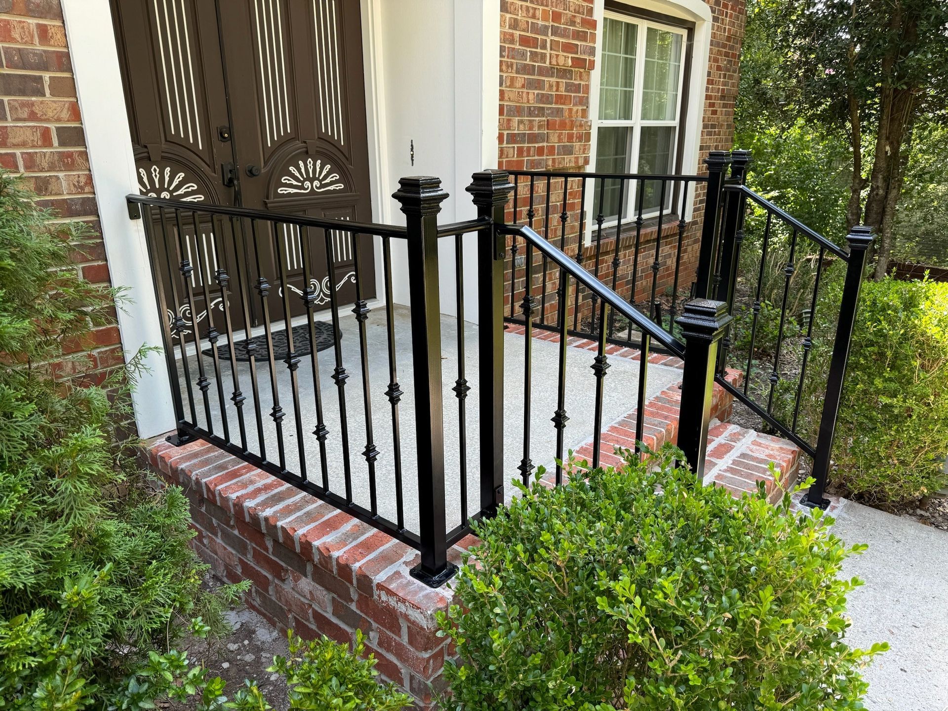 Brick front porch with black wrought-iron railing and double wooden doors, bordered by green shrubs.