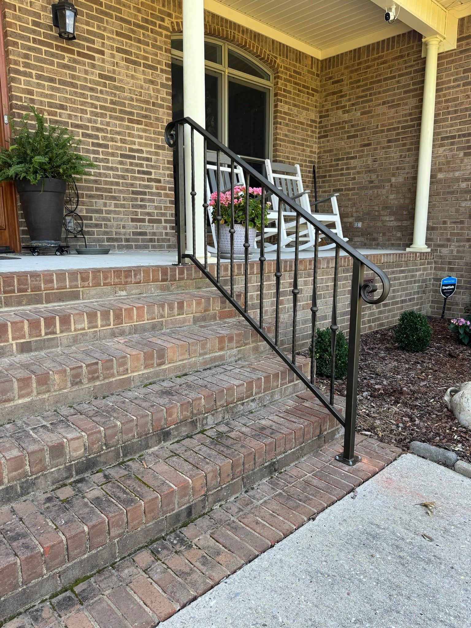 Brick front steps with black metal handrail leading to a house entrance, with potted plants on the porch.