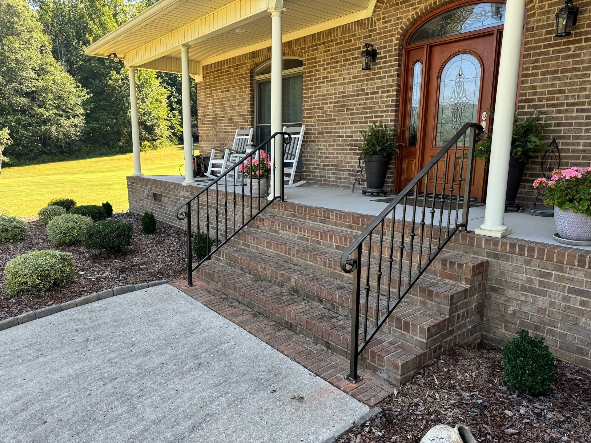 Brick house front porch with steps, black railings, and a concrete walkway in a landscaped yard