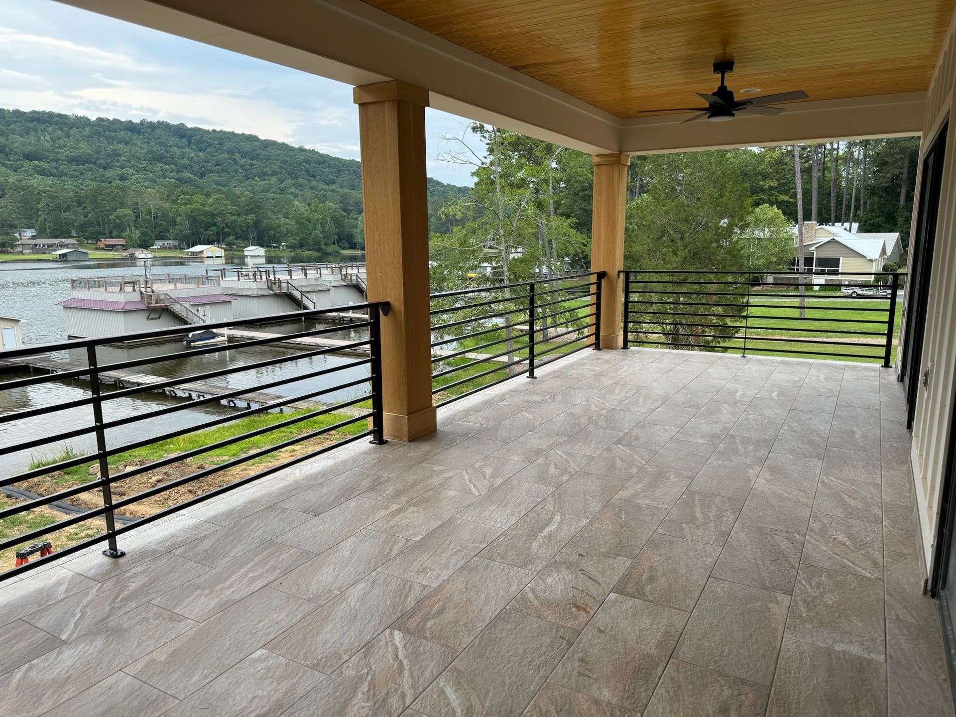 Covered balcony with wood ceiling, black railing, and a lake and forest view beyond