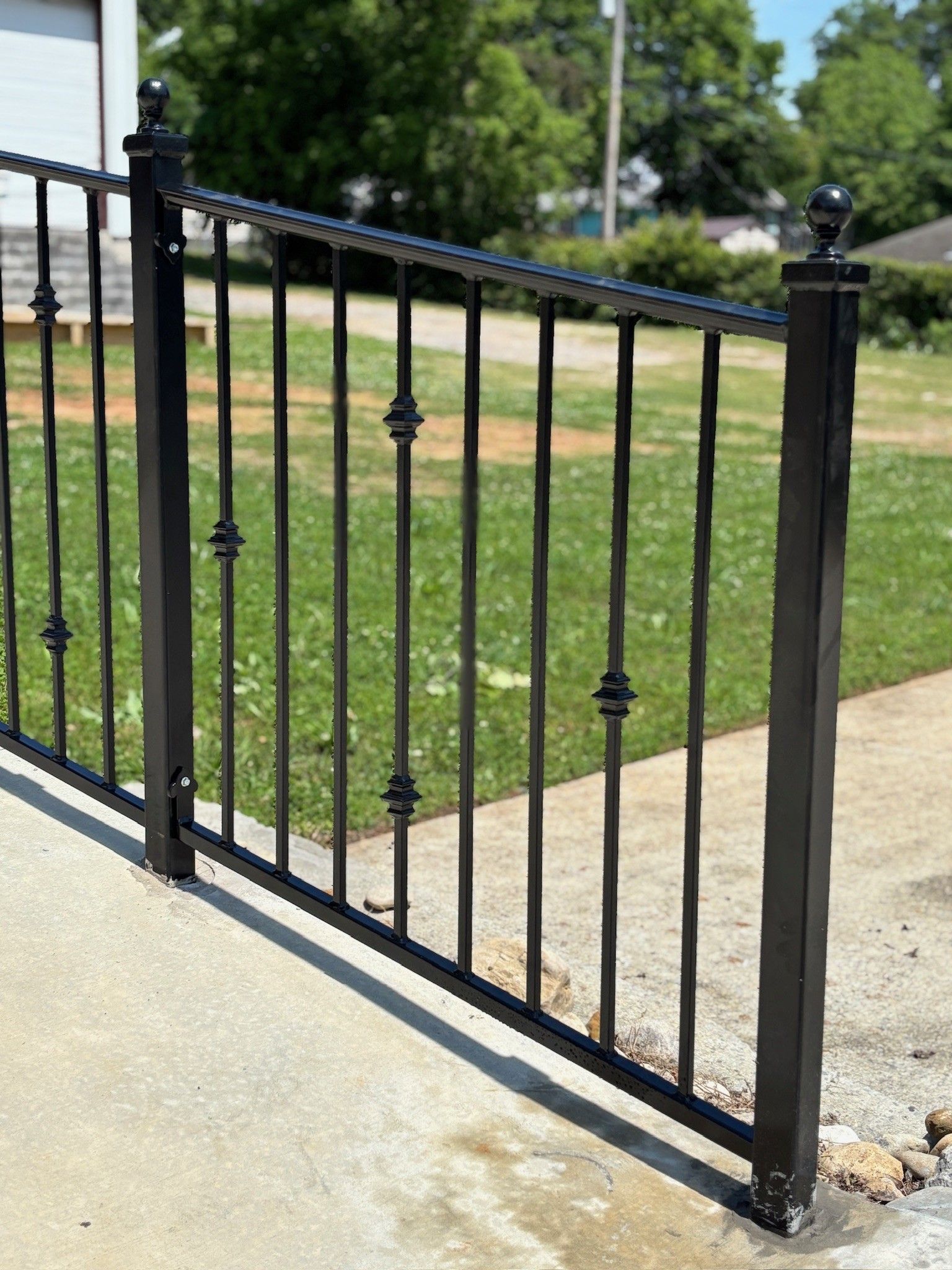 Black metal gate with decorative spindles beside a concrete driveway and grassy yard