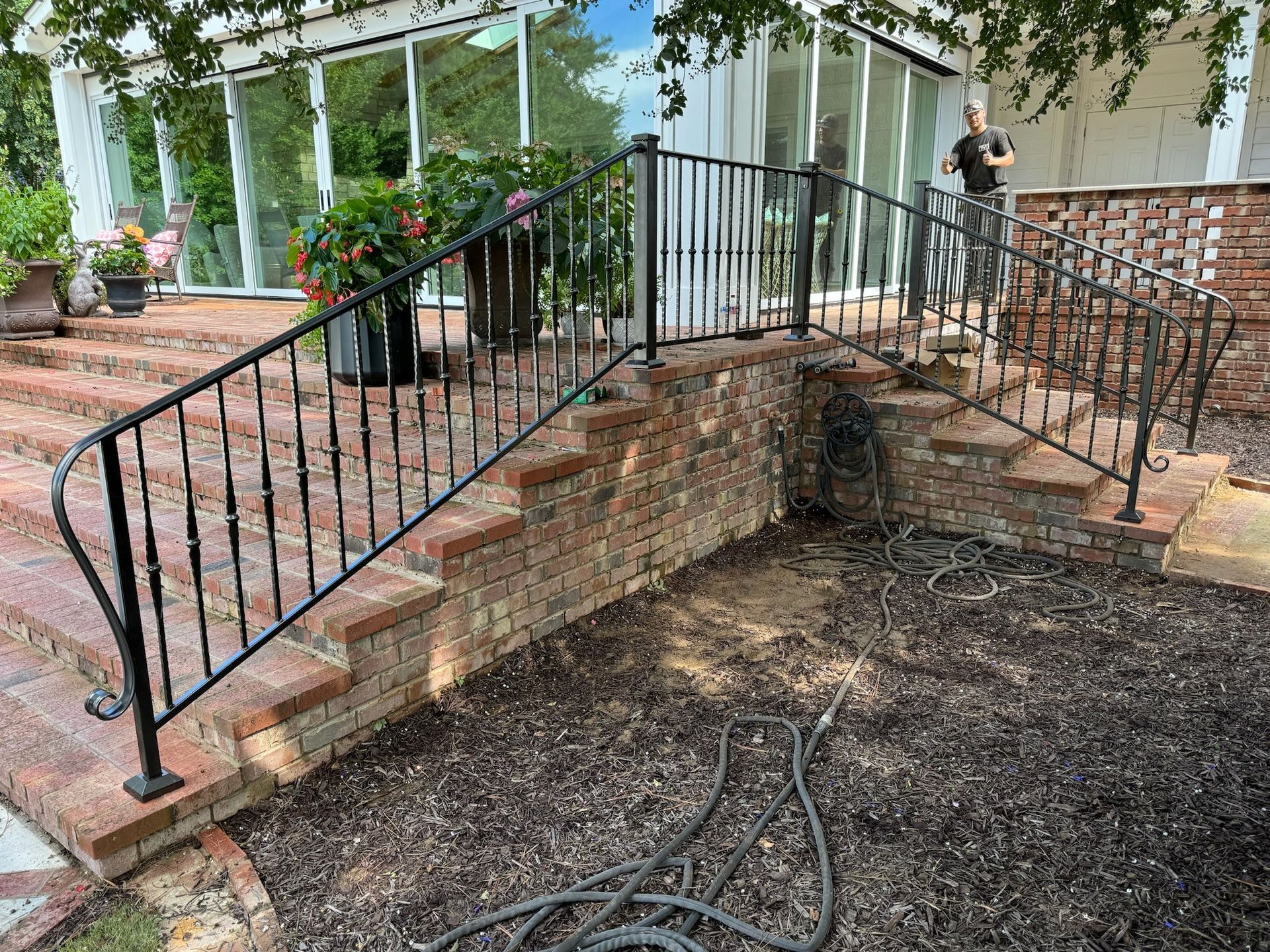 Brick steps with black railings leading to a porch, with a person standing near the doorway and a hose on the ground.