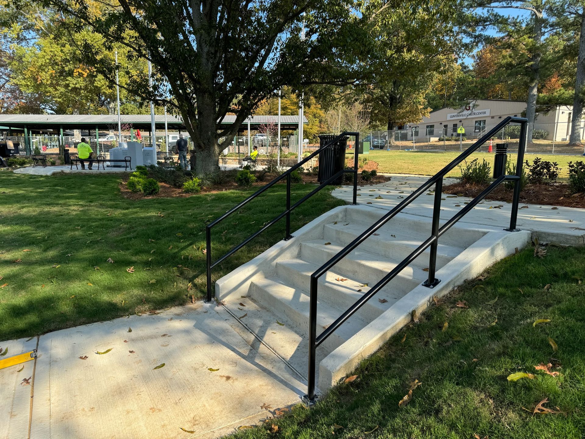Accessible concrete ramp with black railings in a park, with grass, trees, and buildings in the background