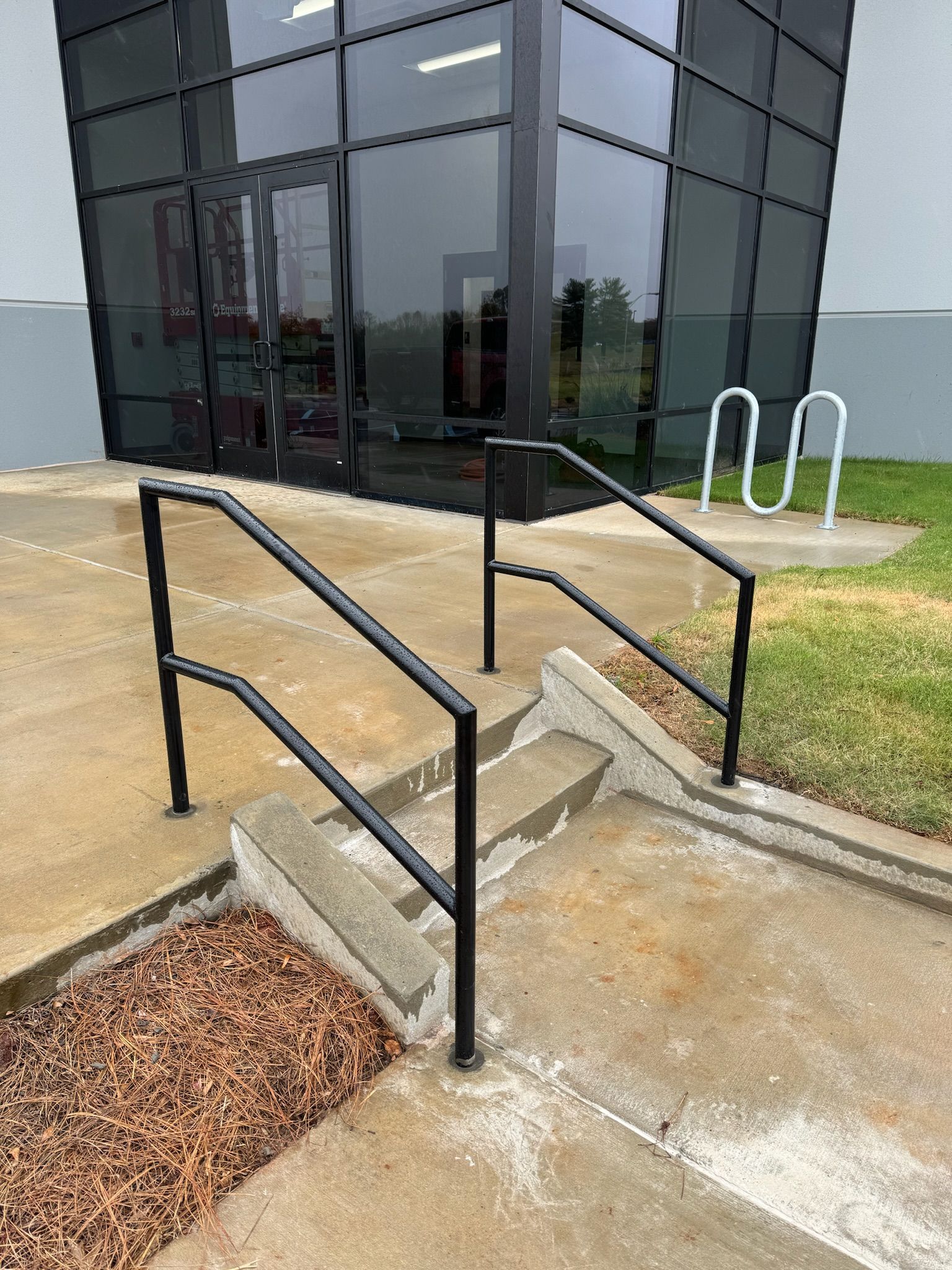 Concrete steps with black handrails leading to a glass building entrance beside a bike rack and mulch bed
