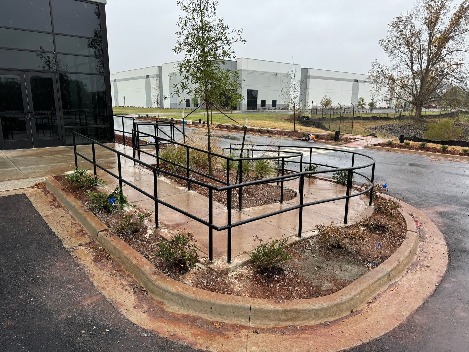 Outdoor concrete planter with black railings beside a wet parking lot and office building