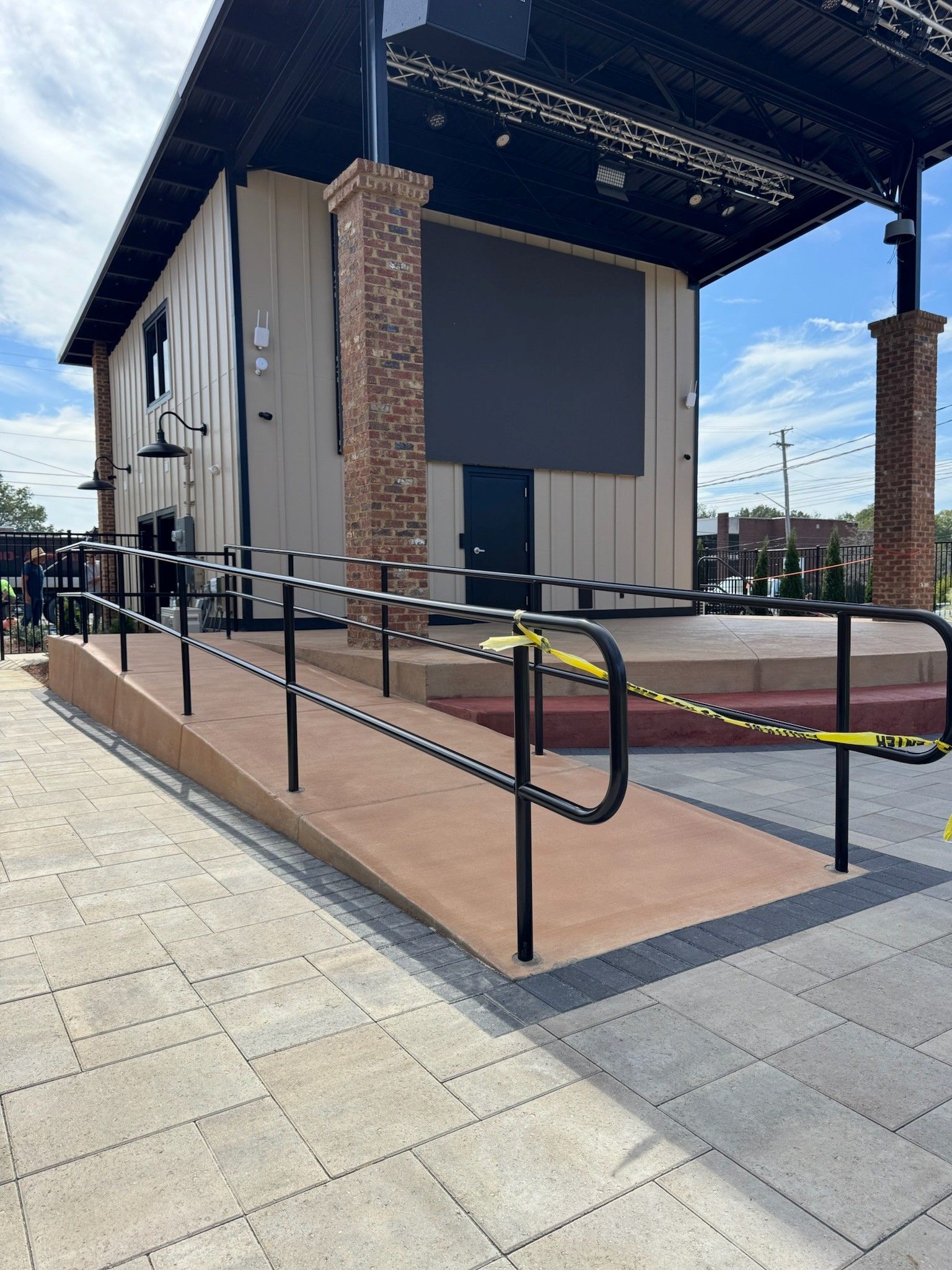 Outdoor stage with black roof, brick columns, and ramps with black railings in a paved plaza