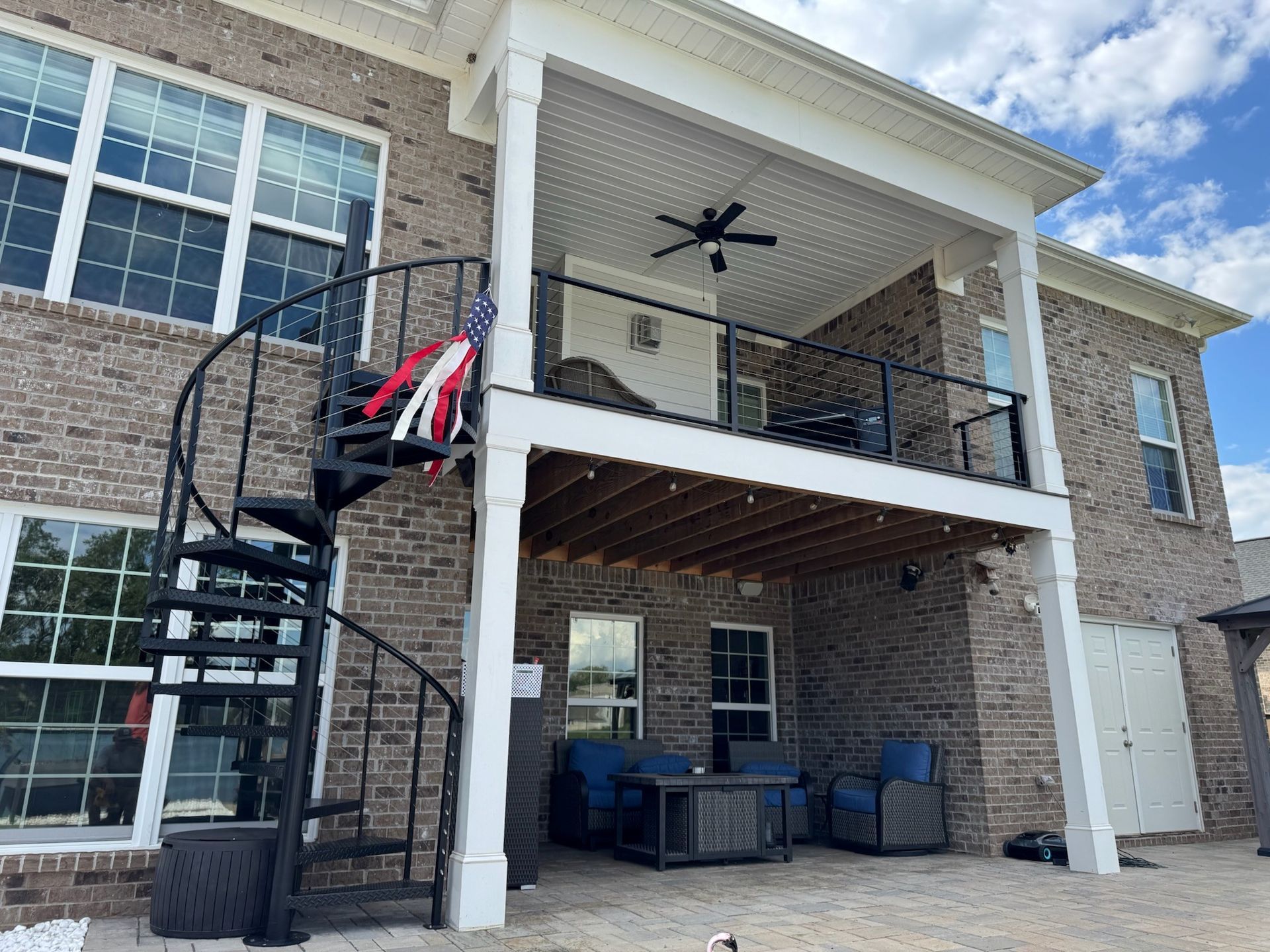 Covered patio beneath a brick house with white columns, spiral stairs, and outdoor seating