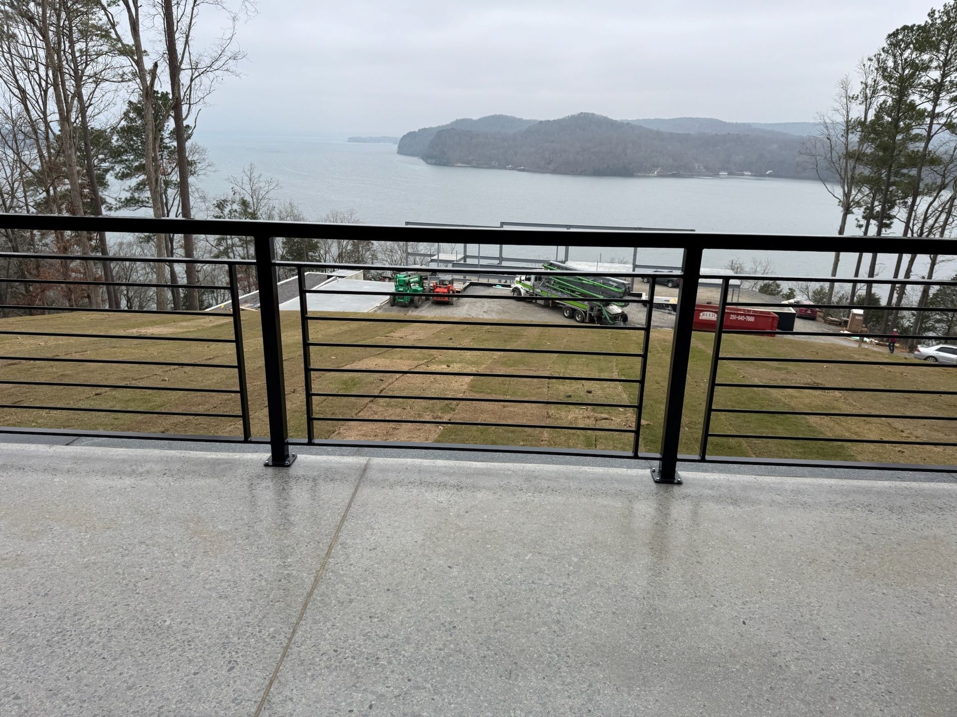Rainy waterfront view from a balcony with black railing, grassy shore, and distant island.