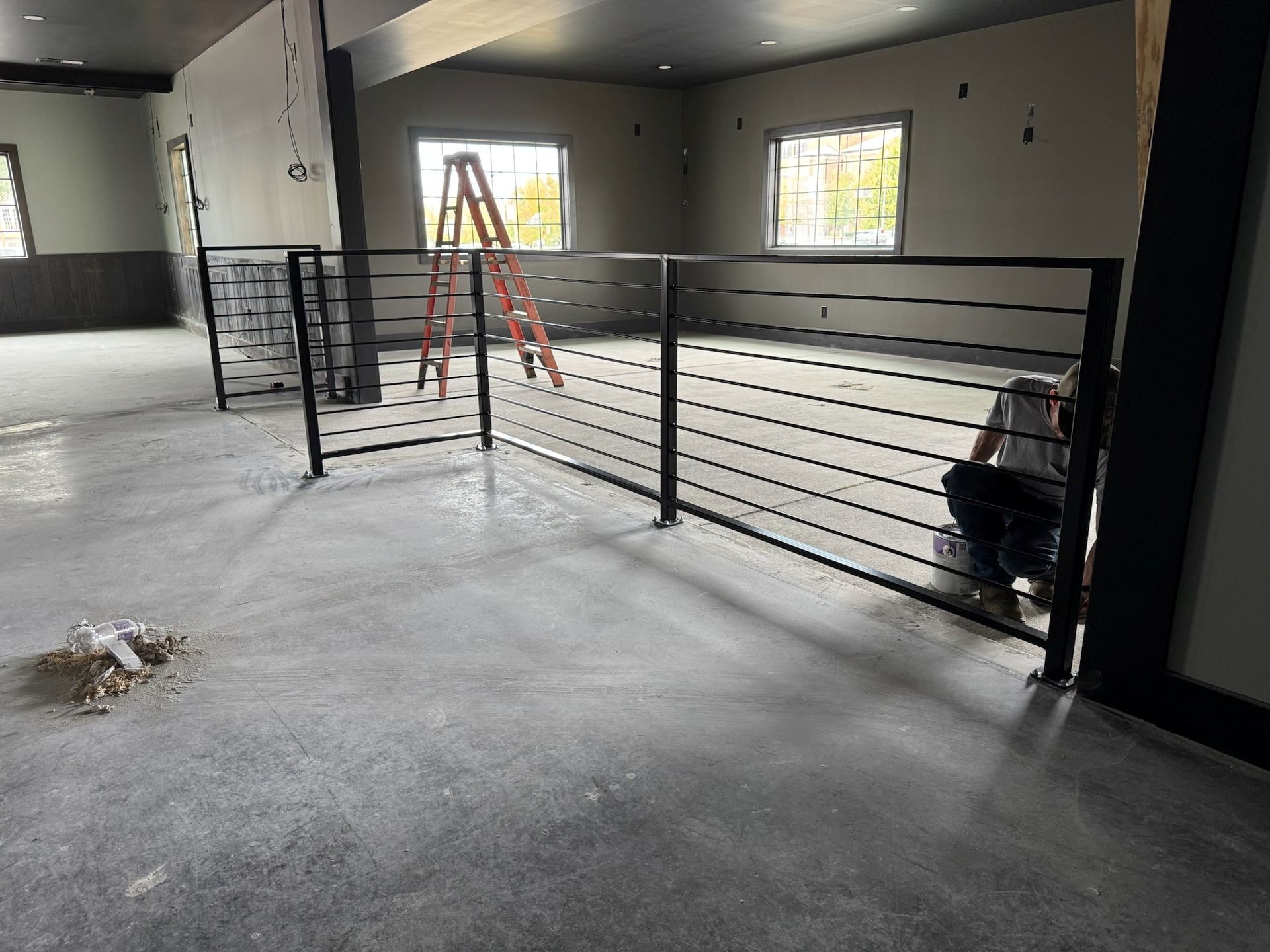 Indoor renovation with gray concrete floor, black railing, ladder, and tools near windows