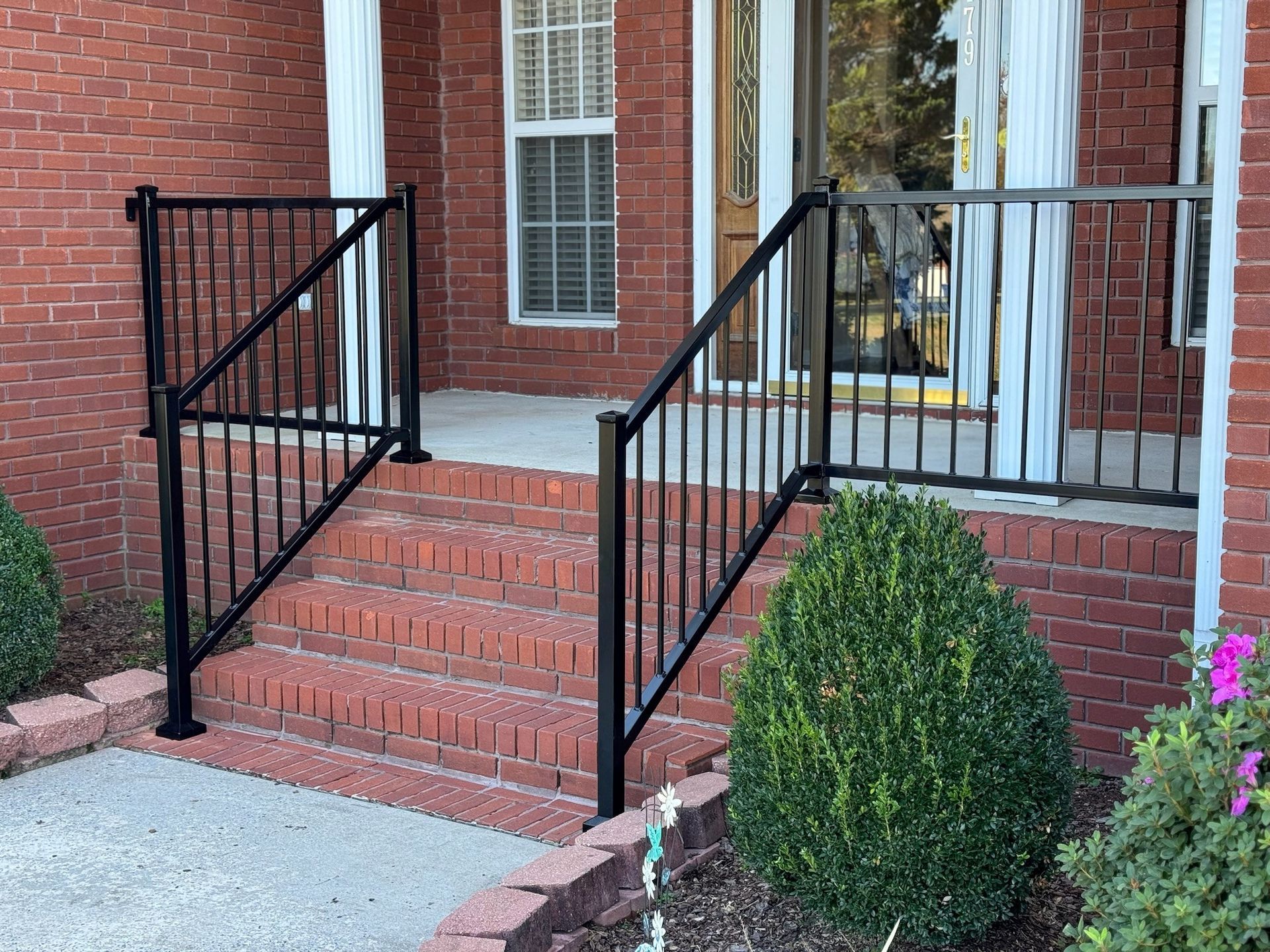 Brick house entrance with red steps, black railings, and a small shrub by the walkway