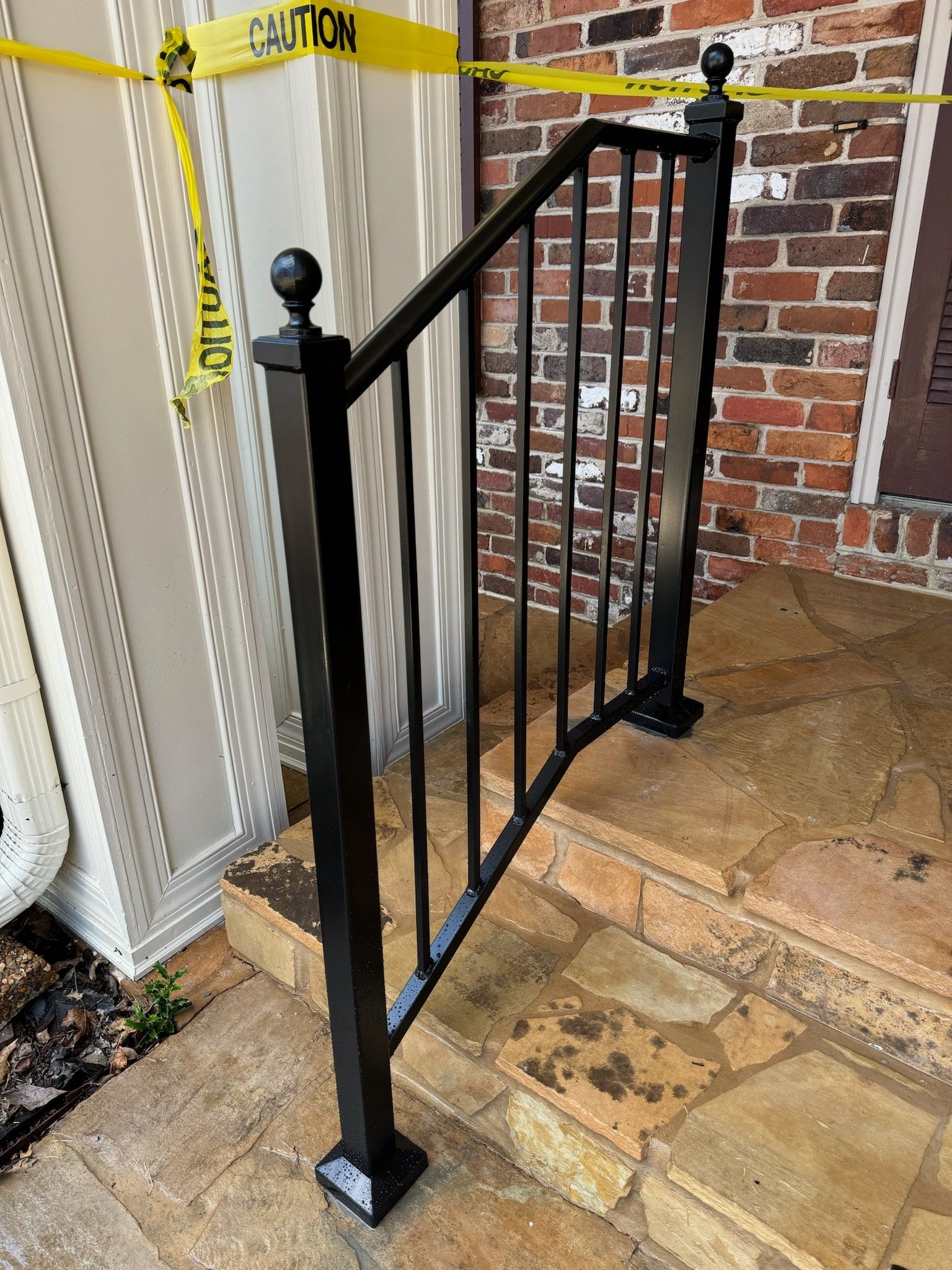 Black metal handrail and gate beside brick steps, with caution tape across the entrance.