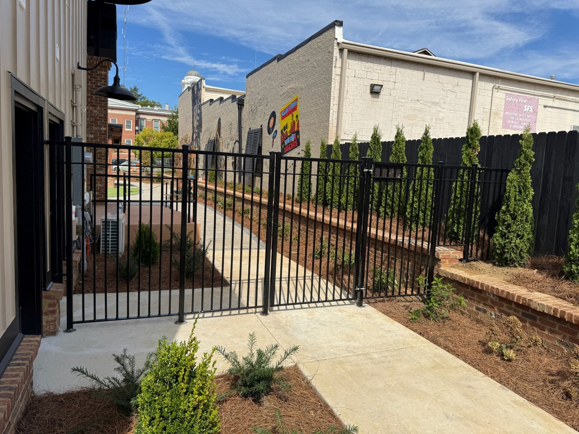 Gated apartment courtyard with a concrete walkway, black fence, and landscaped beds under a blue sky