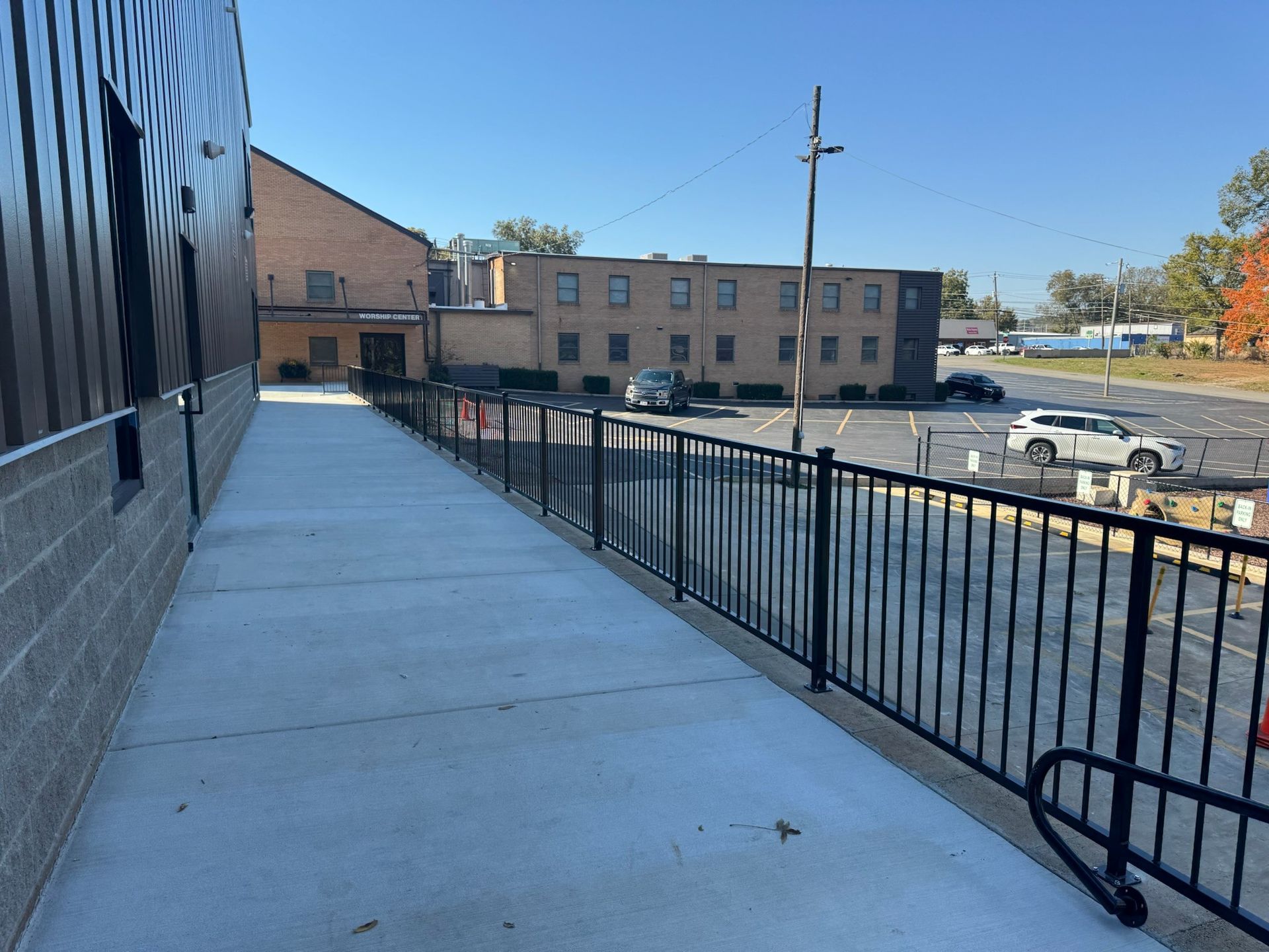 Empty concrete walkway beside a black metal railing, overlooking a parking lot and beige building on a sunny day