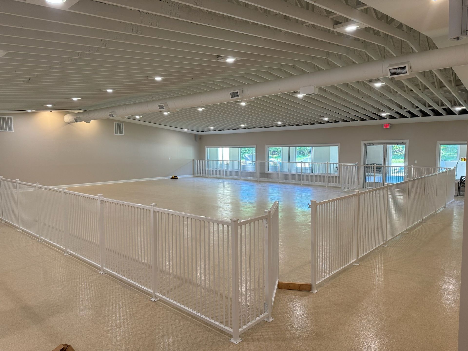 Large empty indoor sports court with white fencing, high ceiling, and windows along the far wall