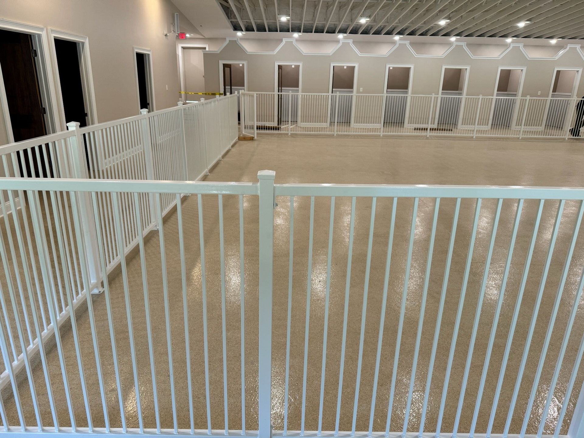 Empty indoor dog kennel with white gates and beige floors, viewed from behind a white railing.