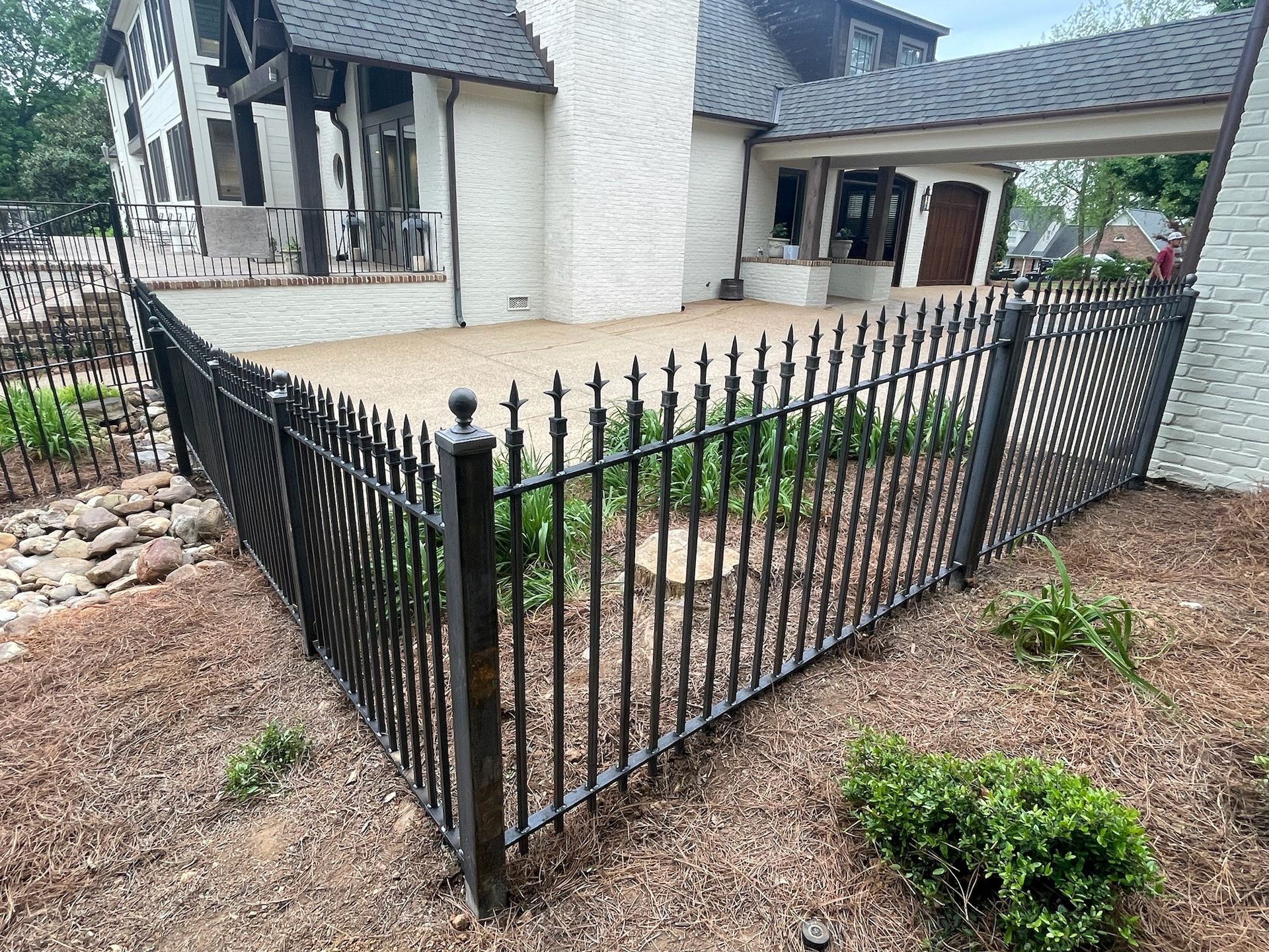 Black metal fence around a concrete patio beside a white house and gravel yard.