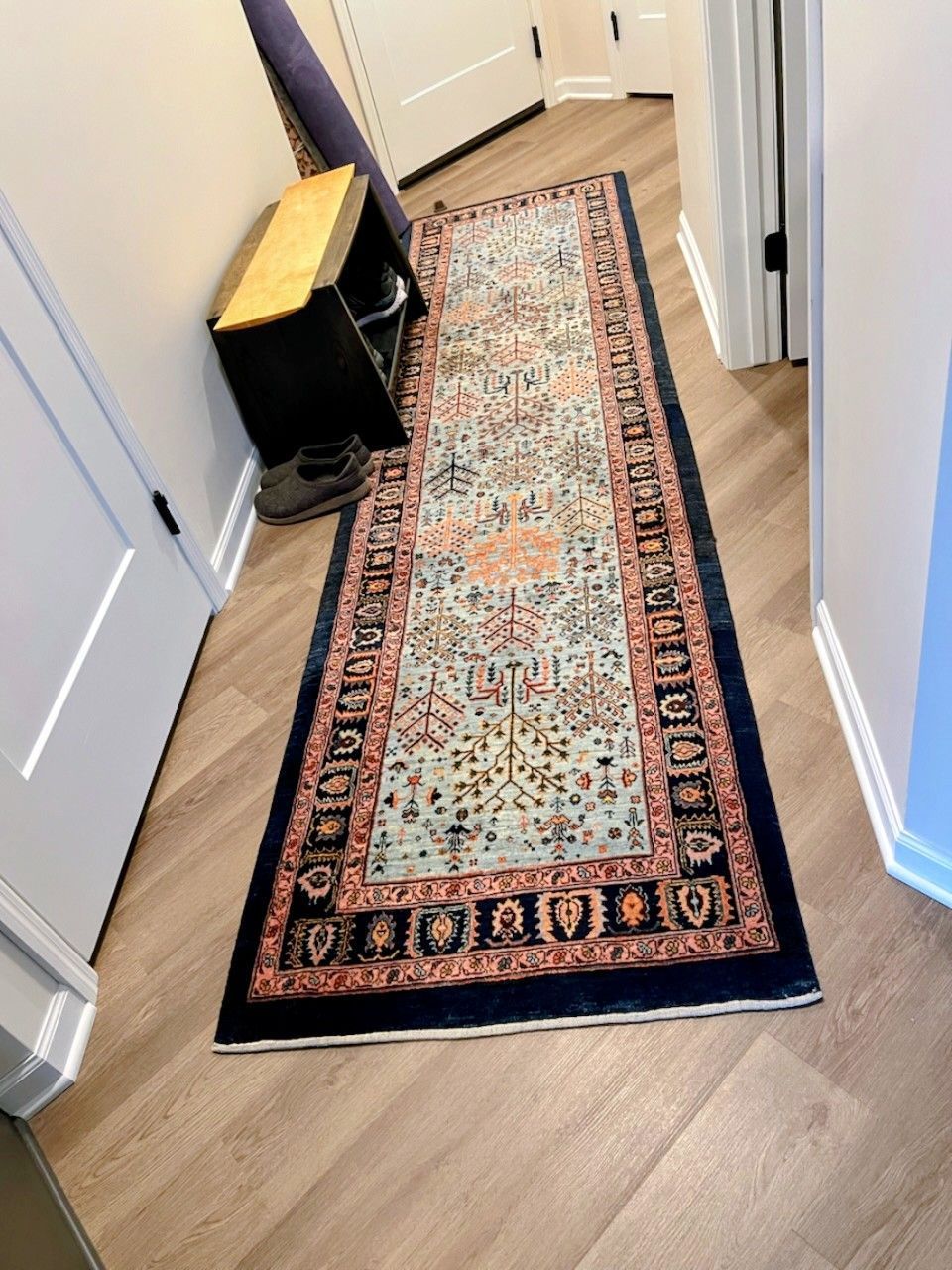 Long hallway with wood-look floor; a blue, coral, and cream runner rug; black shoe shelf; and closed white doors.